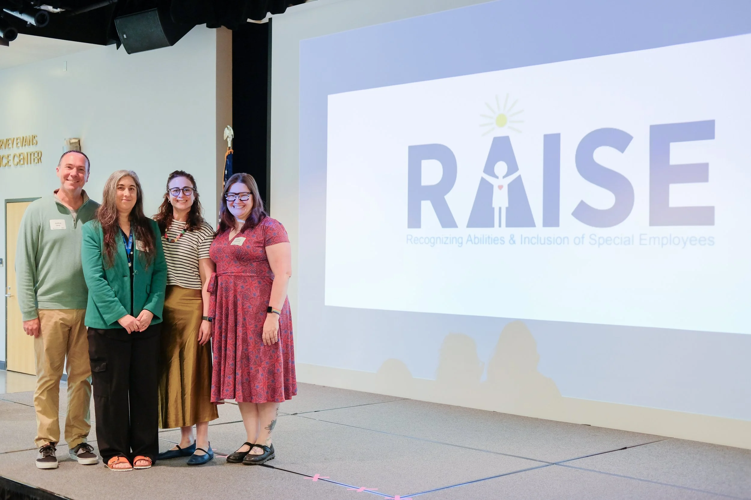 Four professionally dressed people stand on a stage, smiling at the camera, during the RAISE graduation ceremony.