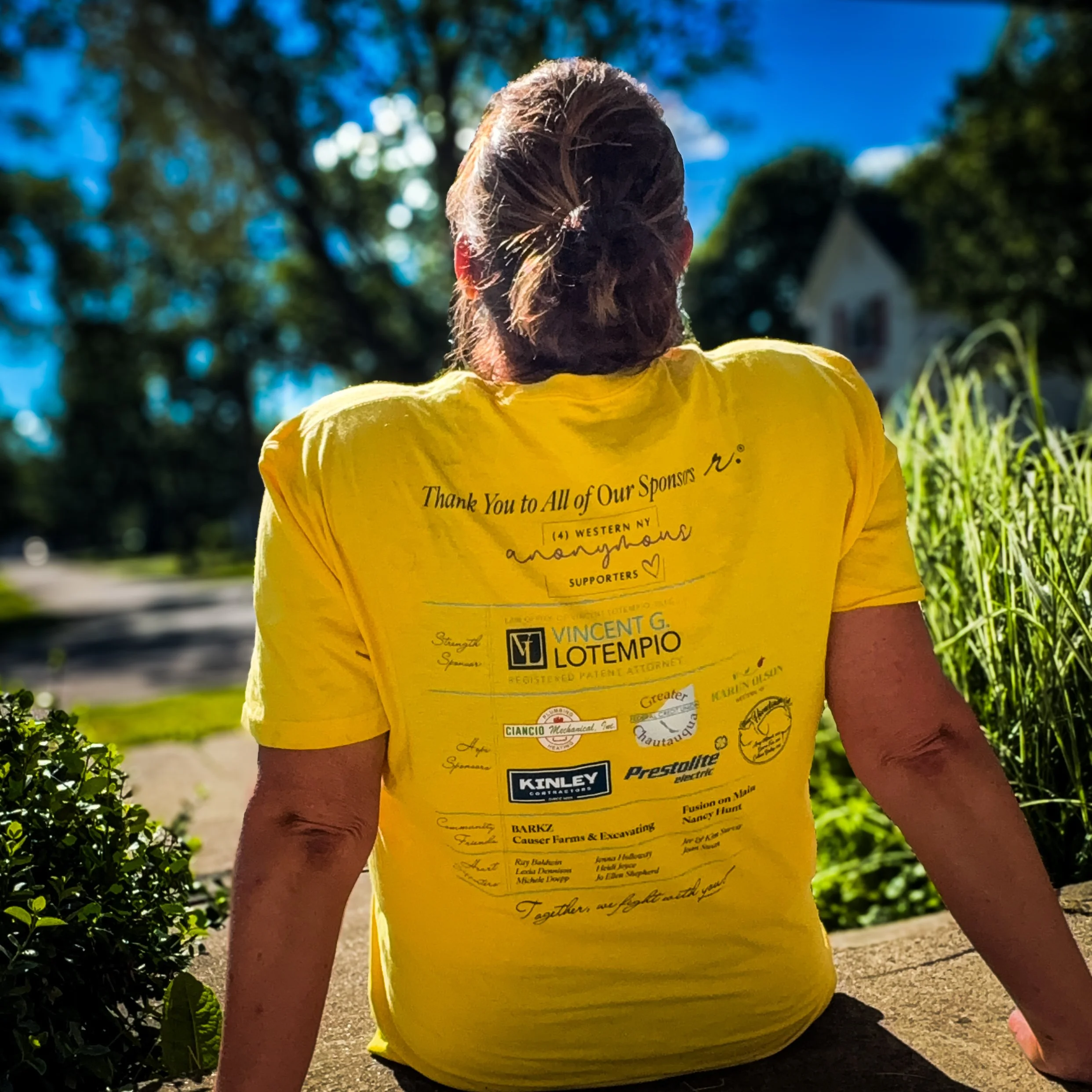 Kim Survil wearing a yellow t-shirt that displays all of the sponsors on the back on a sunny evening before the donation day.