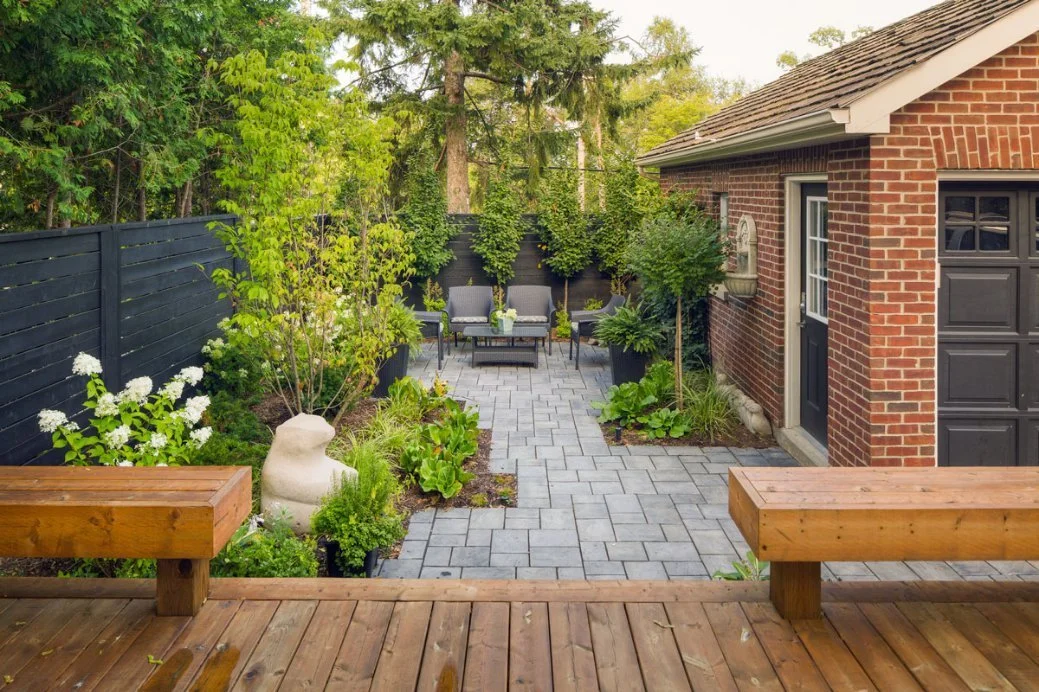Backyard patio with paver stones, outdoor seating, lush green plants, trees, and a brick house with a black garage door.