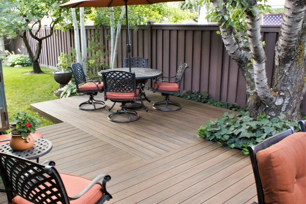 Wooden backyard patio with a round table and four chairs, a large tree, potted plants, green grass, and a brown fence.