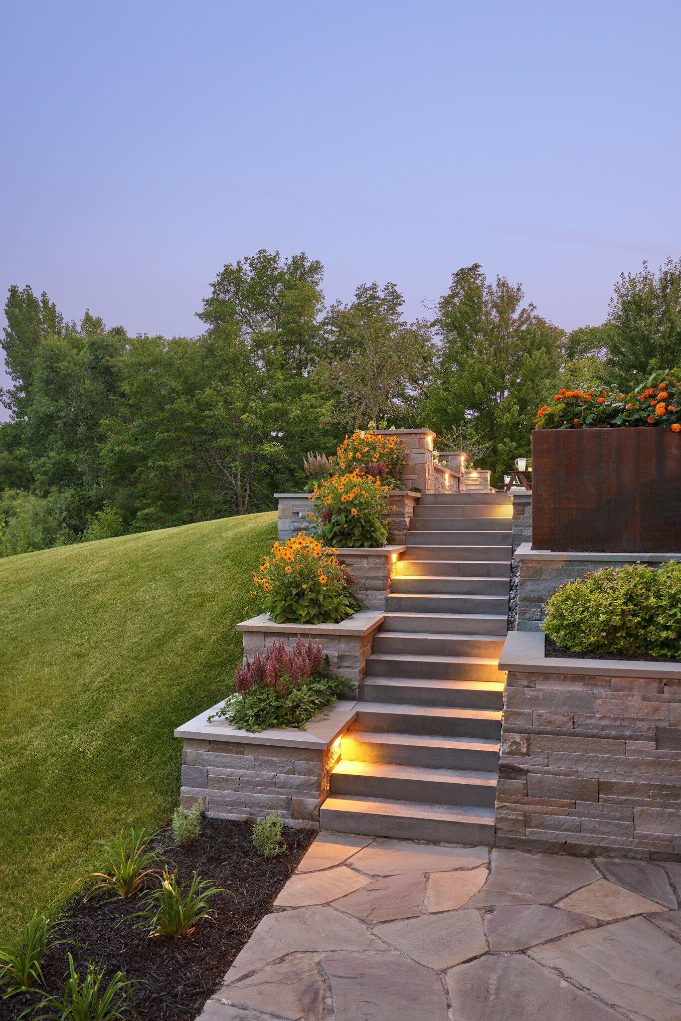 Outdoor landscaped steps with built-in lighting, flower planters, and stone pathway, with trees in the background during dusk.