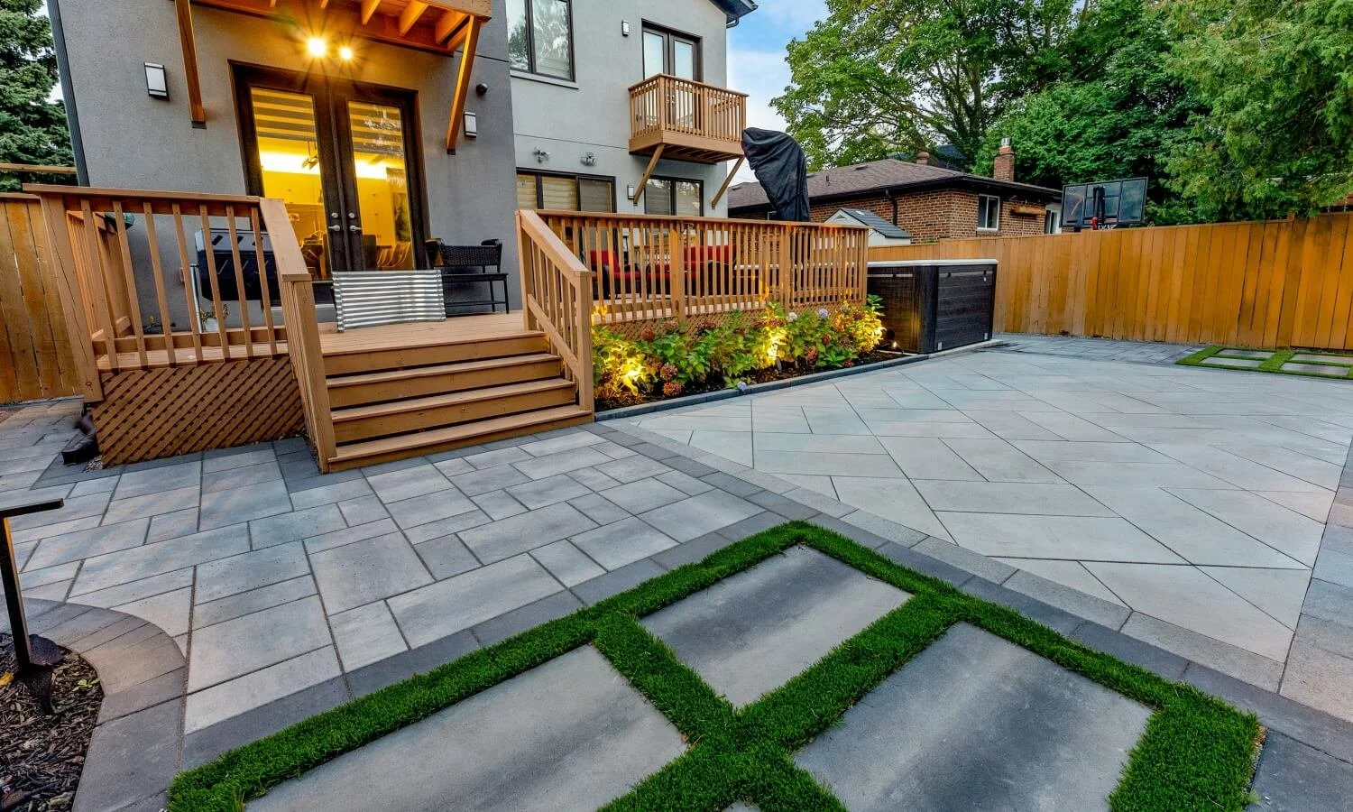 Backyard patio with stone tiles, a wooden deck with stairs, a flower bed with lighting, a fence, and a basketball hoop in the background.