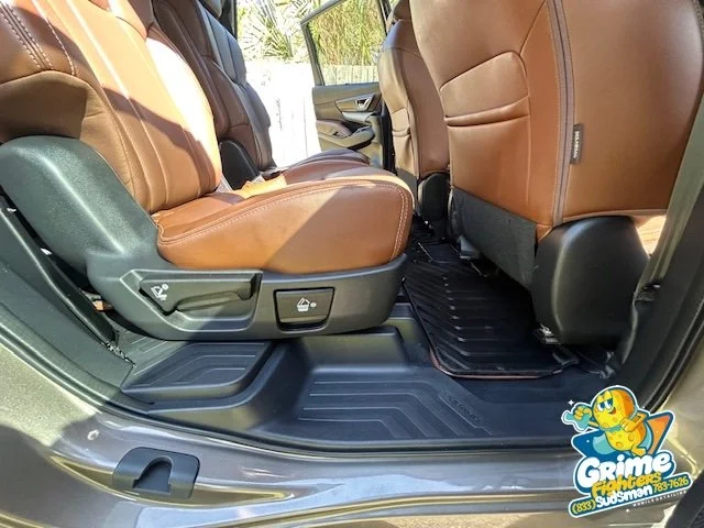 Inside view of a vehicle's rear passenger area, showing brown leather seats, black floor mats, and a partly open rear door with sunlight filtering in.