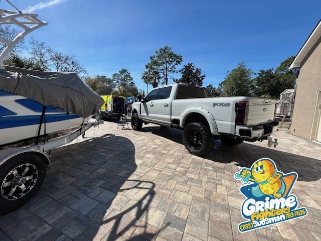 A white pickup truck parked on a curved brick driveway with boats and equipment in the background, and trees under a partly cloudy sky.