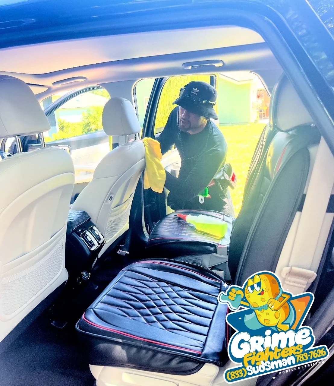 A person cleaning the interior of a car with a yellow cloth, wearing a black hat and black clothing, with cleaning supplies on the back seat, showing the back of a white car.