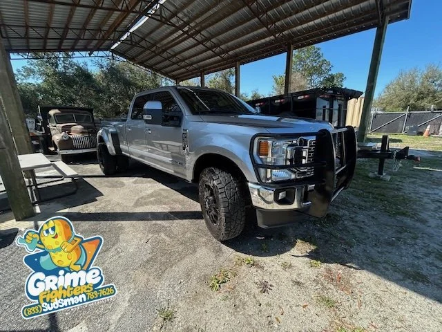 Silver and black lifted pickup truck parked under a metal roofed carport, with two other vintage vehicles parked adjacent.