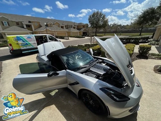 A silver sports car with its hood open and driver's door ajar parked in a residential area. A white maintenance van is parked nearby, and there are townhouses with a manicured lawn and bushes in the background under a partly cloudy sky.
