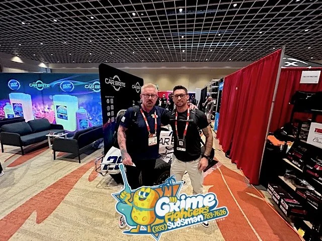 Two men standing at a trade show booth with banners and red partition panels, holding a promotional sign for Crime Fighters.