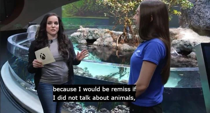 Xbox host interviewing a Seattle Aquarium staff member in front of a live marine exhibit during the Xbox Earth Day Twitch stream, and an on-screen caption reading "because I would be remiss if I did not talk about animals"