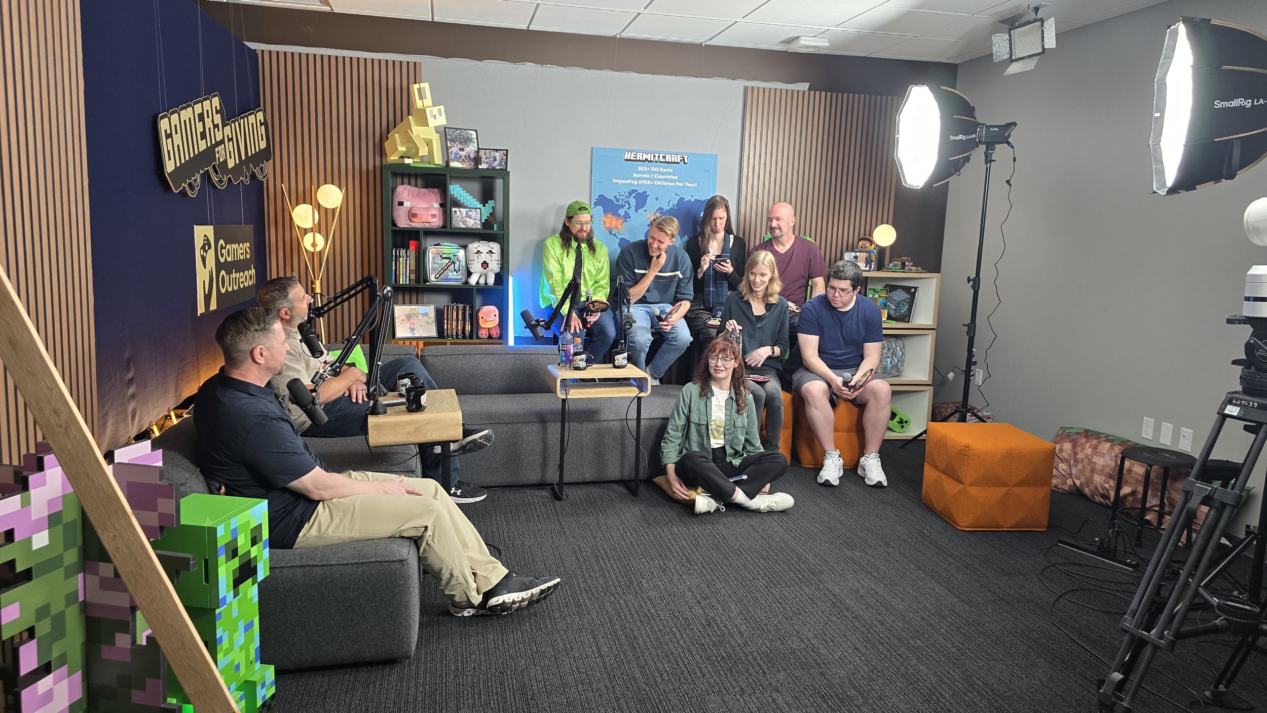 A group of gaming community members gathered at Mojang's office during a Hermitcraft and Gamers for Giving event, featuring Gamers Outreach branding, Minecraft-themed decor, and a Hermitcraft partnership display during a panel recording.
