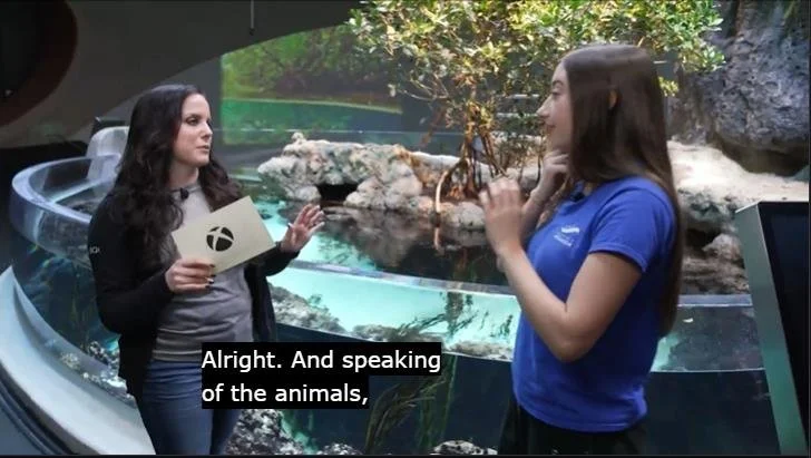 Xbox host interviewing a Seattle Aquarium staff member in front of a live marine exhibit during the Xbox Earth Day Twitch stream, with an on screen caption reading "Alright. And speaking of the animals,"
