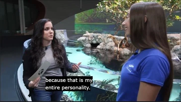 Xbox host speaking with a Seattle Aquarium staff member in front of a marine exhibit, with the on-screen caption reading "because that is my entire personality," during the Xbox Earth Day Twitch stream