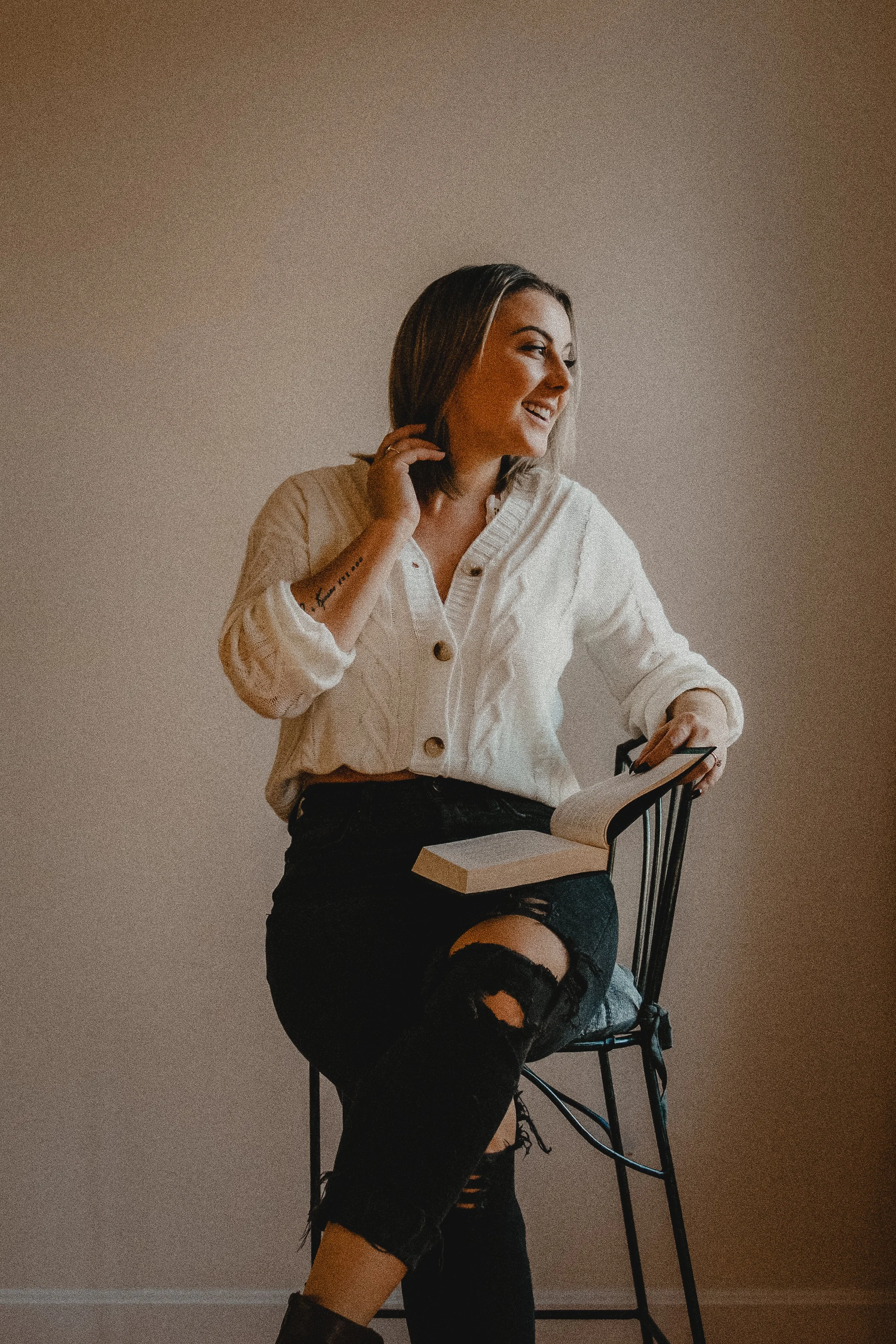 A woman with shoulder-length brown hair, wearing a white knit cardigan and ripped black jeans, sitting on a black chair with a book open on her lap, smiling and looking to her right.