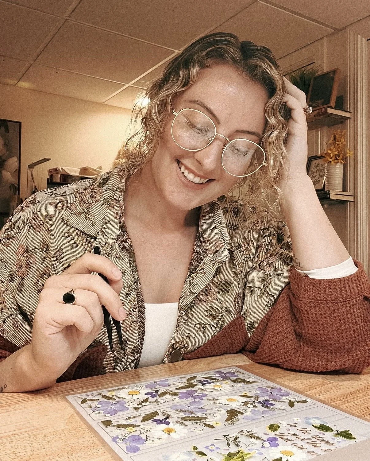 A woman with curly blonde hair, wearing glasses and a floral shirt, is smiling while looking down at a puzzle of pressed flowers on a wooden table.