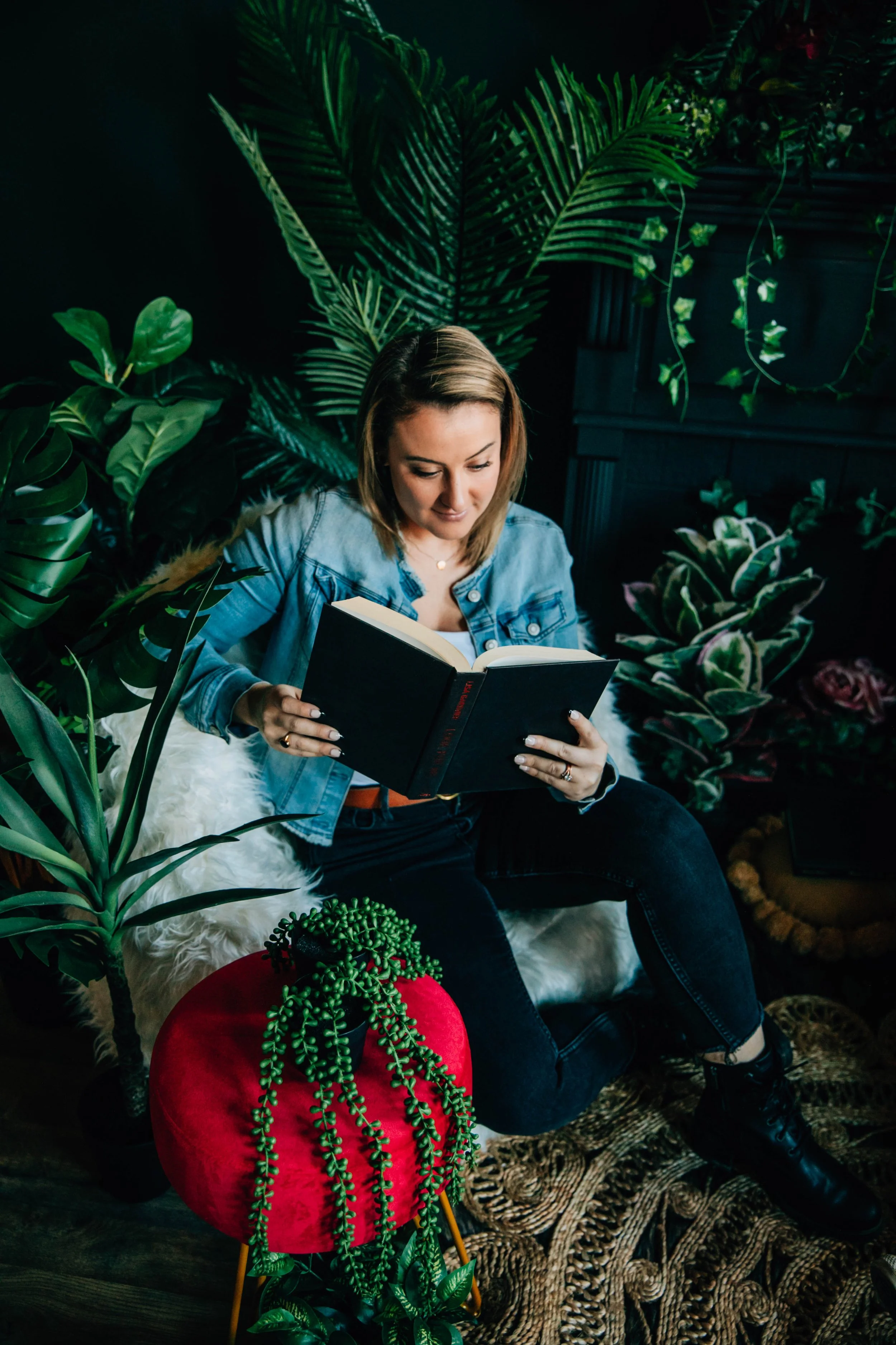 A woman with shoulder-length brown hair sitting on a white furry cushion, reading a black hardcover book, surrounded by green plants in a dark room with black walls.
