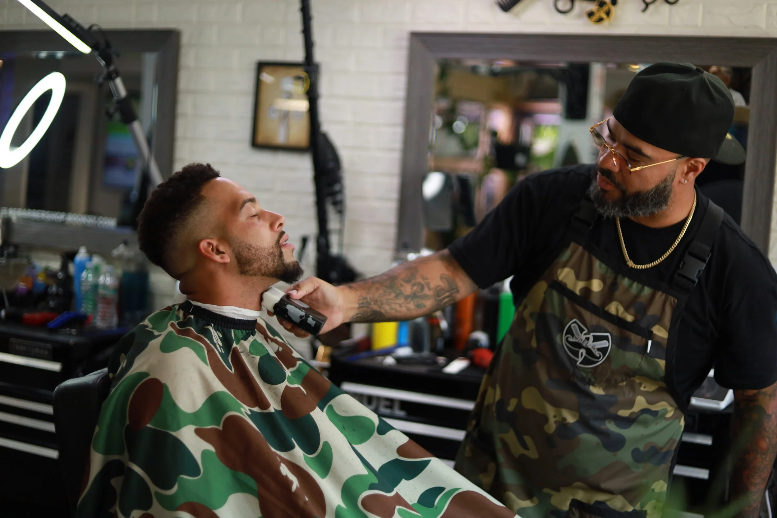 Barber shaving a man's beard in a barbershop while the man sits with a camouflage cape on.