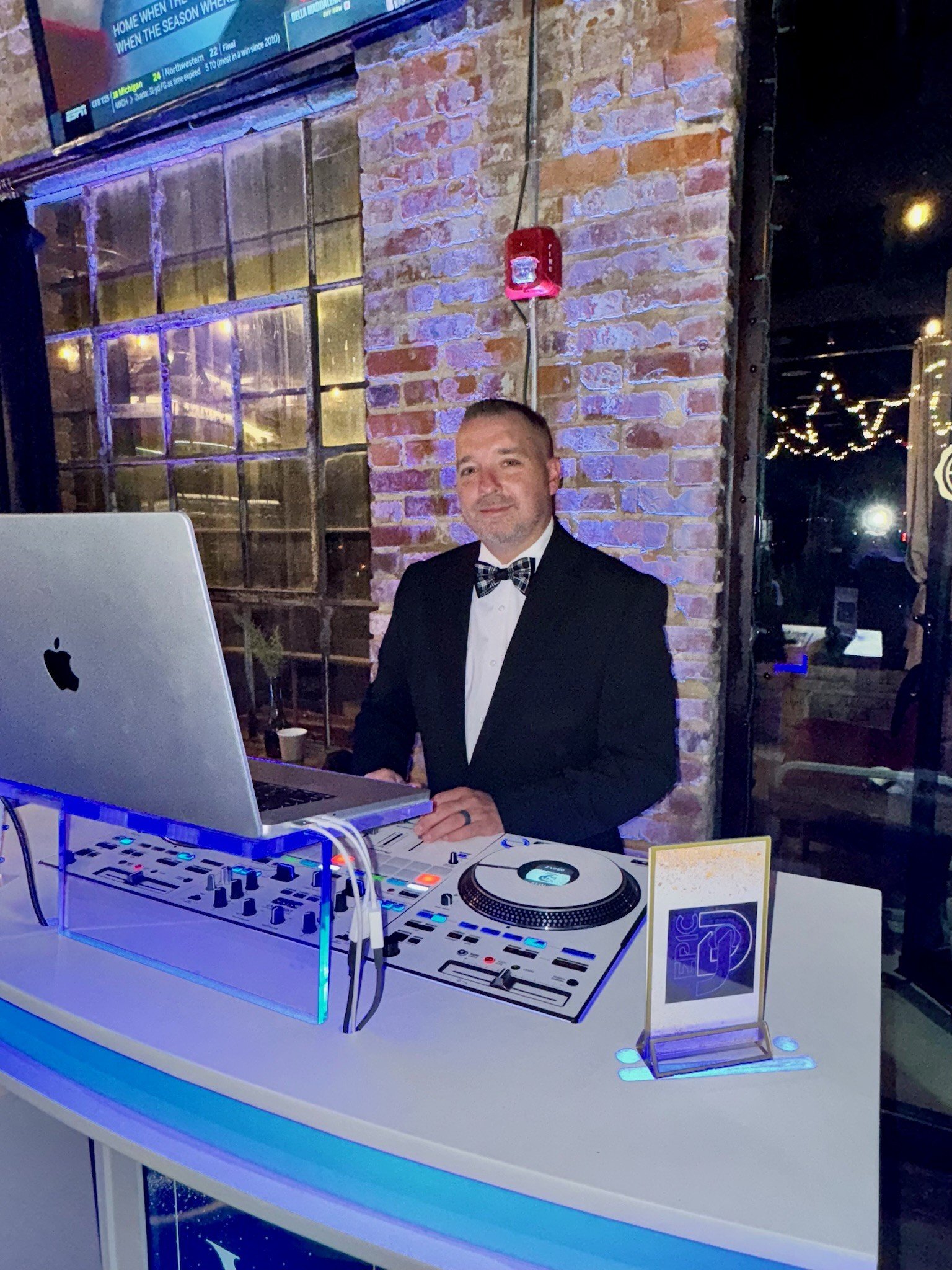 A man dressed in a tuxedo with a bow tie standing behind a DJ console with a laptop and turntable, in a venue with a brick wall and large windows, at night.