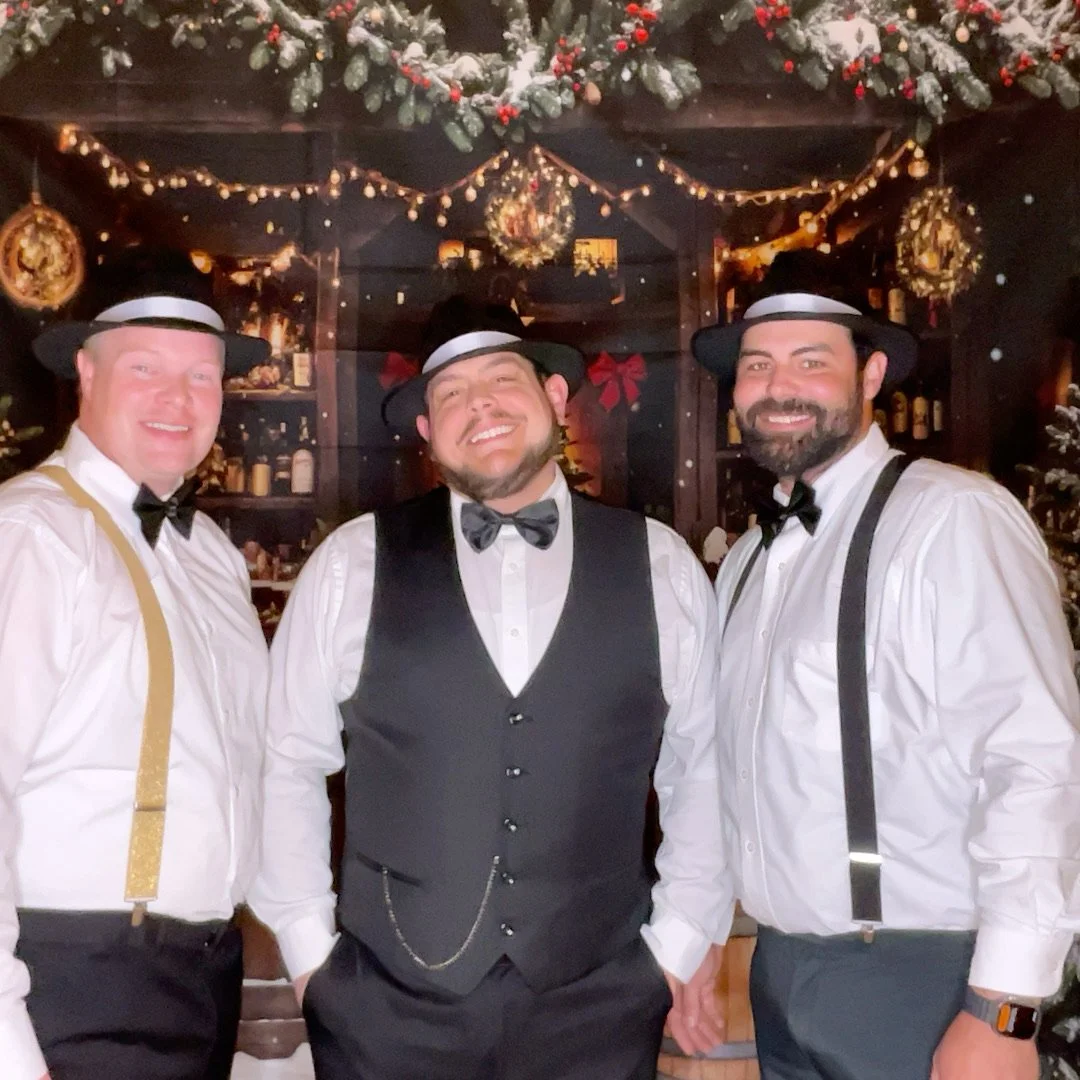 Three men dressed in vintage style with white shirts, black suspenders, bow ties, and black hats, smiling in front of a Christmas-themed background with garlands and wreaths.