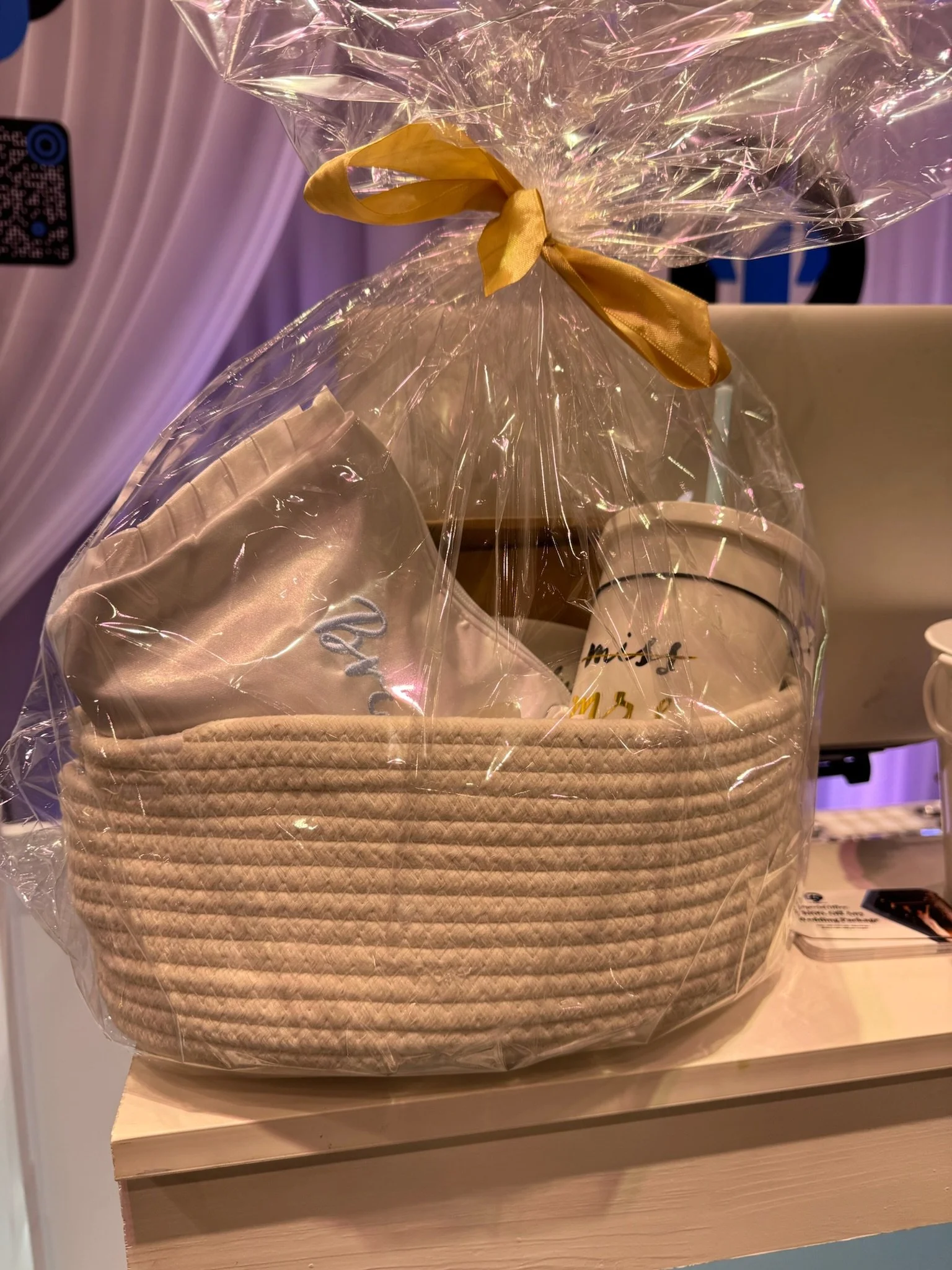 Gift basket wrapped in clear cellophane with a tan ribbon, containing embroidered fabric, ceramic mug, and other items.