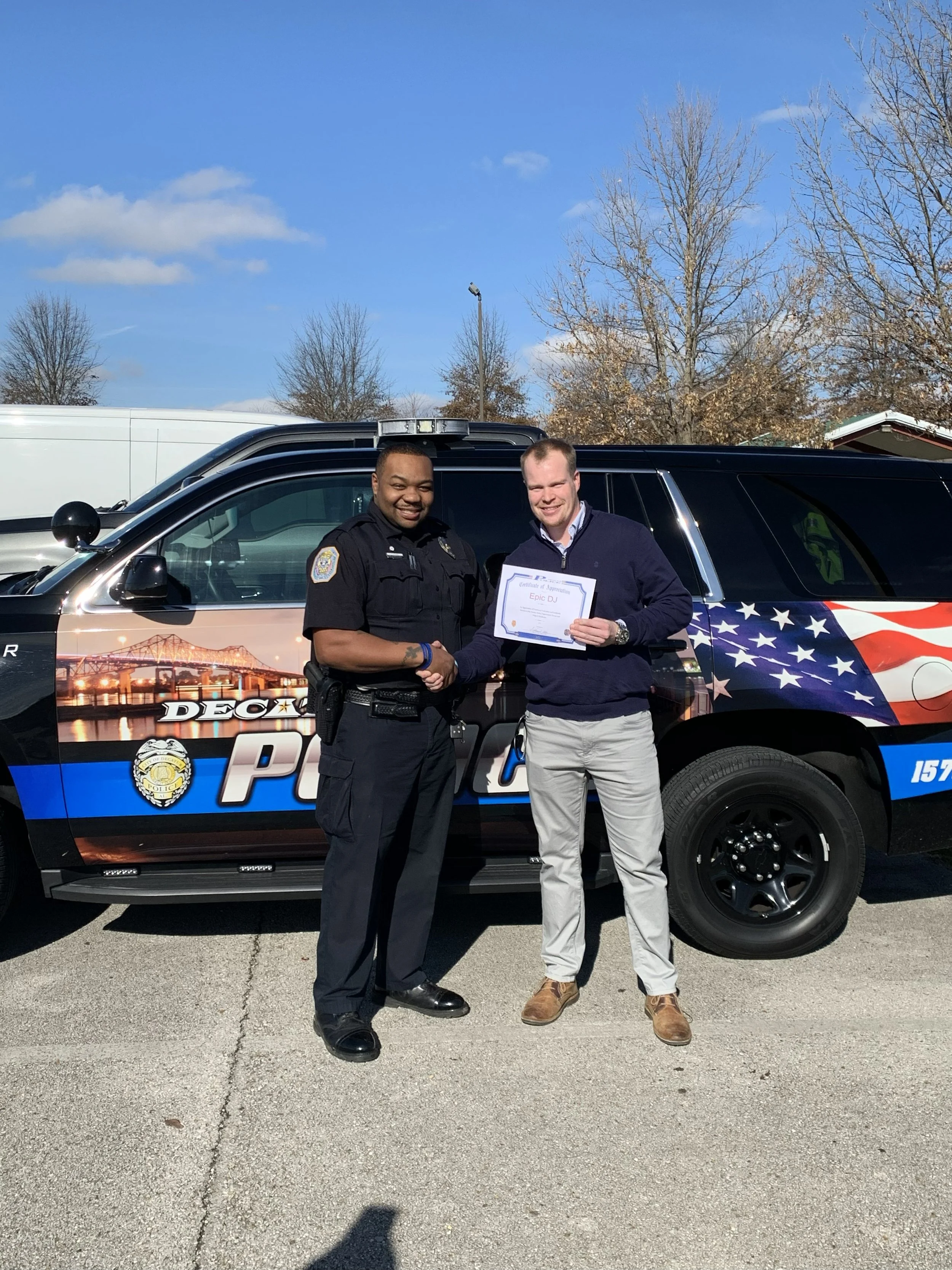 A man in a police uniform shaking hands with a man holding a certificate in front of a police vehicle with a patriotic design and American flag. Clear sky and leafless trees in the background.