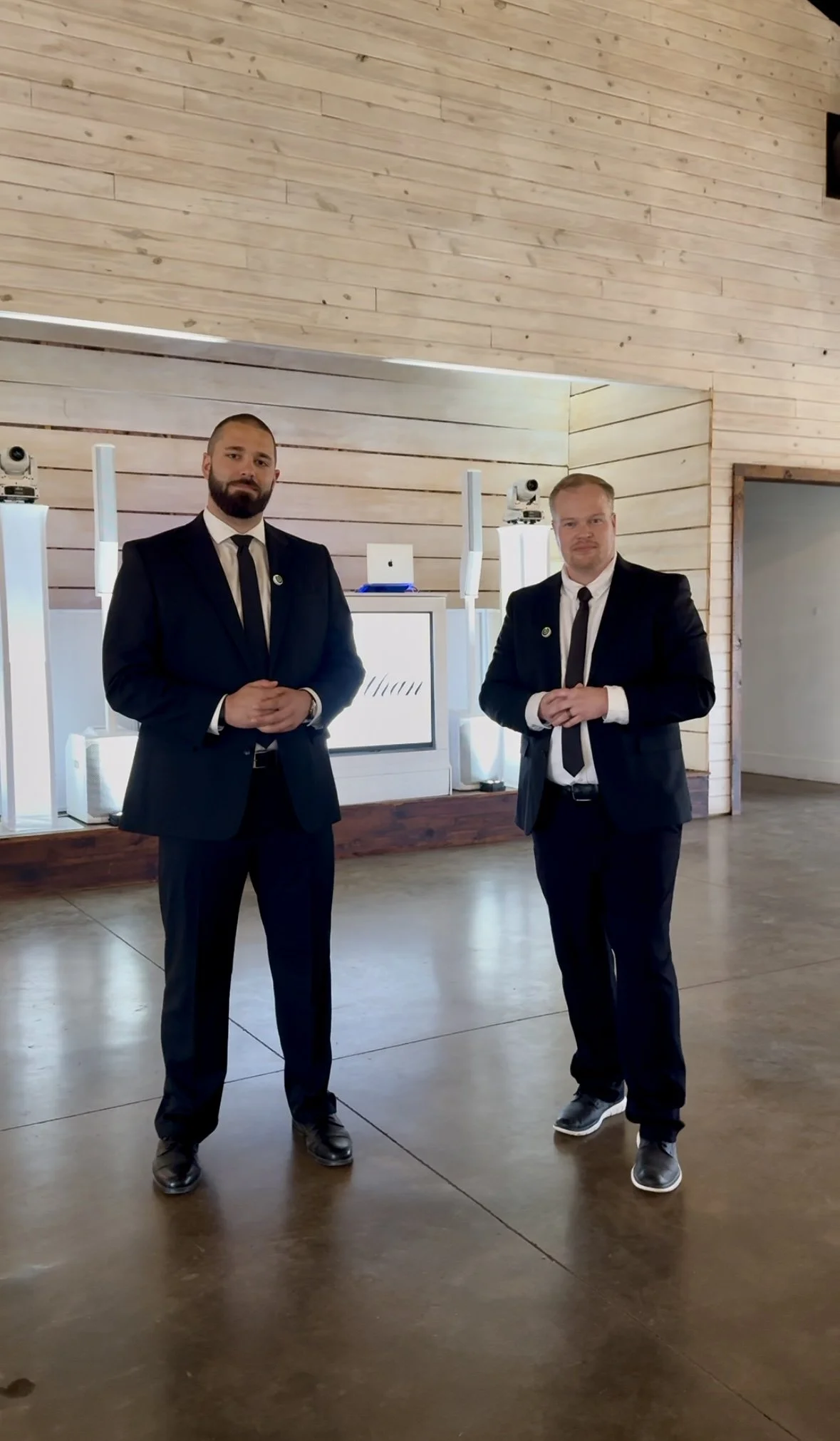 Two men in black suits standing indoors with wood-paneled walls and a bar with a computer and cameras in the background.