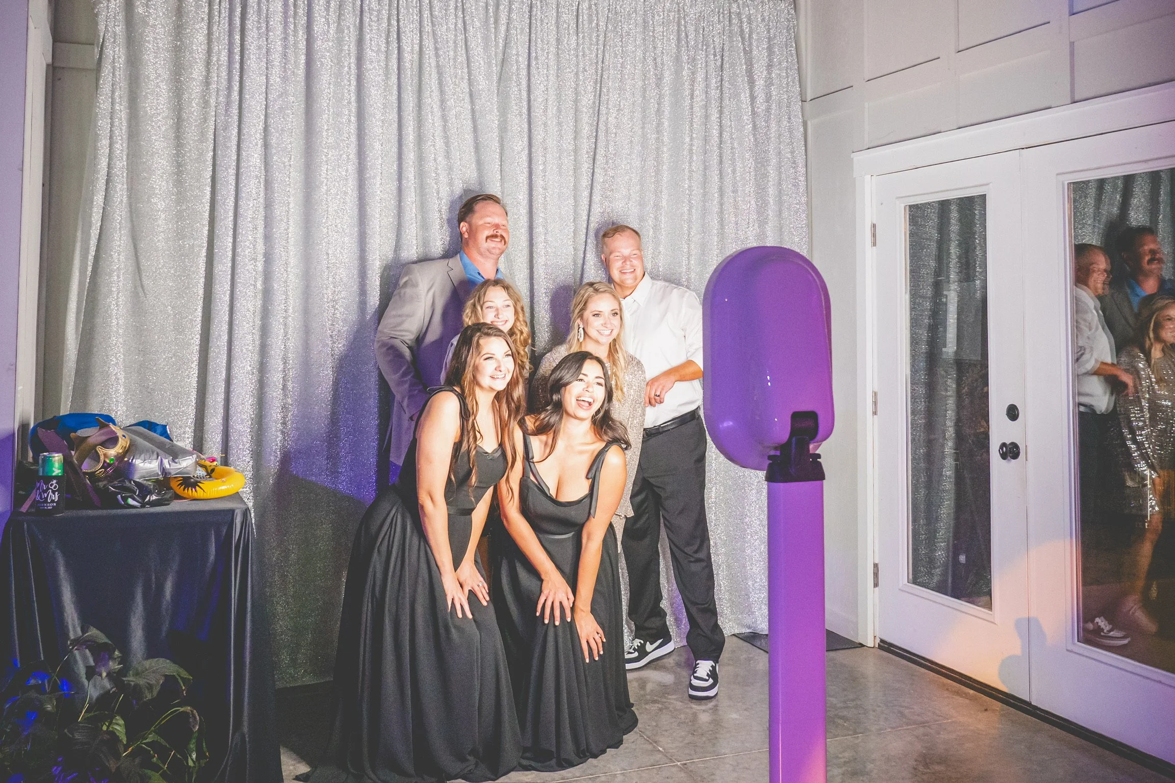 Group of six people taking a fun photo at a celebration event, standing in front of a silver, glittery backdrop with a photo booth camera in the foreground.