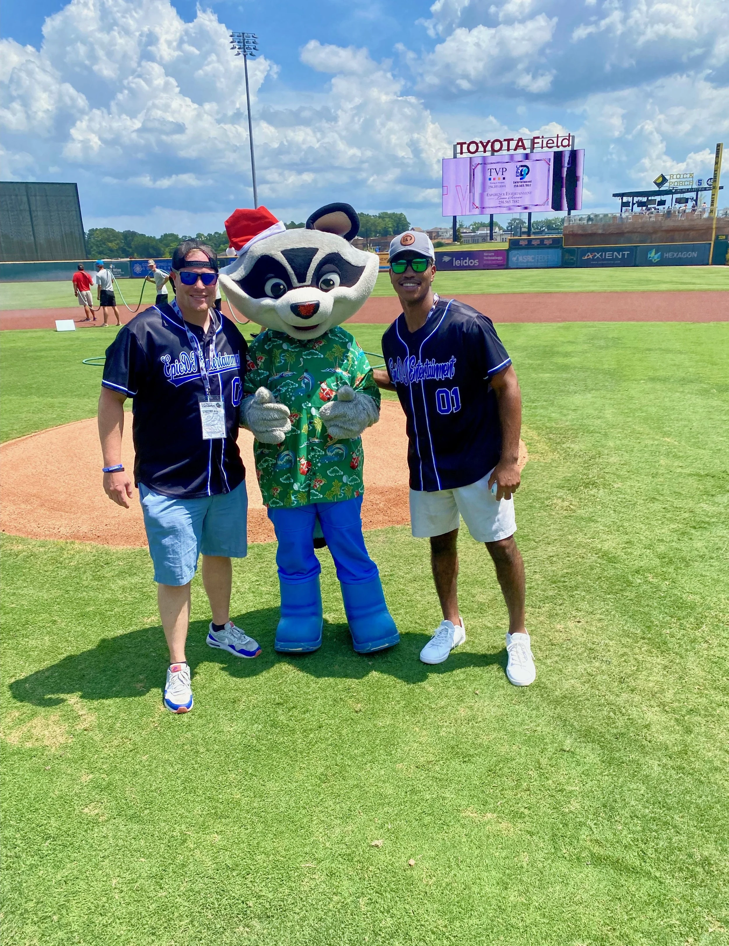 Two men in baseball jerseys standing with a person in a raccoon costume wearing a Hawaiian shirt and a Santa hat at a baseball field during daytime.