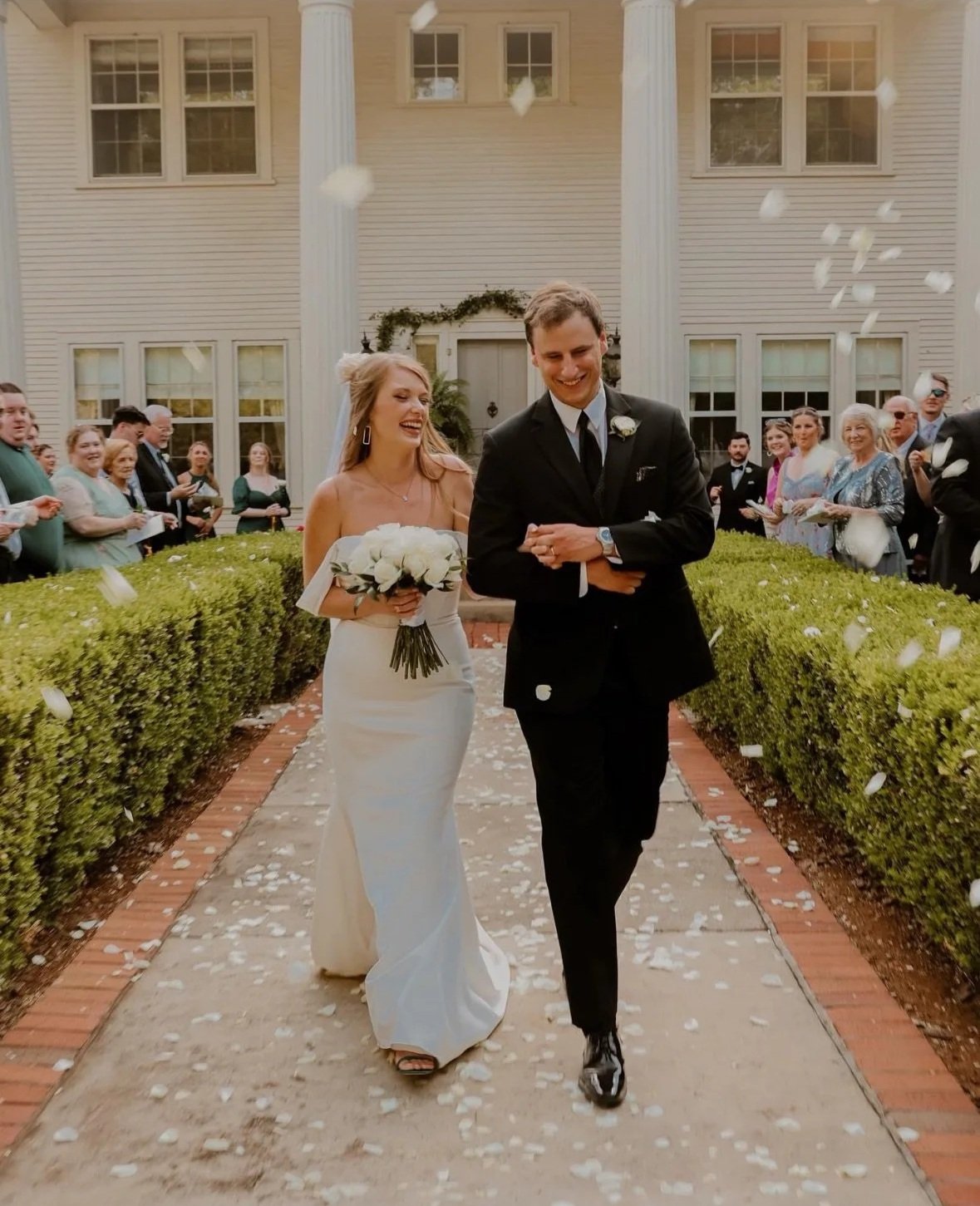 A bride and groom walk down the aisle at their wedding ceremony, surrounded by guests, with white flower petals falling around them.