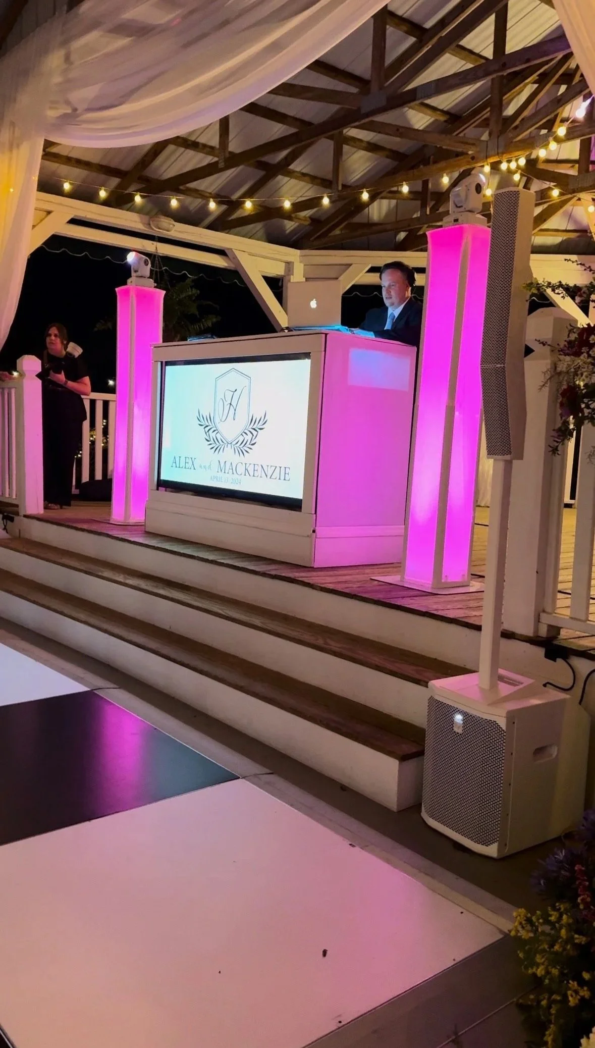 A DJ on a raised outdoor stage with pink lighting, wearing a dark suit, using a laptop and DJ equipment at a wedding reception. A screen displays "Alex and Mackenzie" with a date, April 2024. There are string lights overhead and a woman standing to t