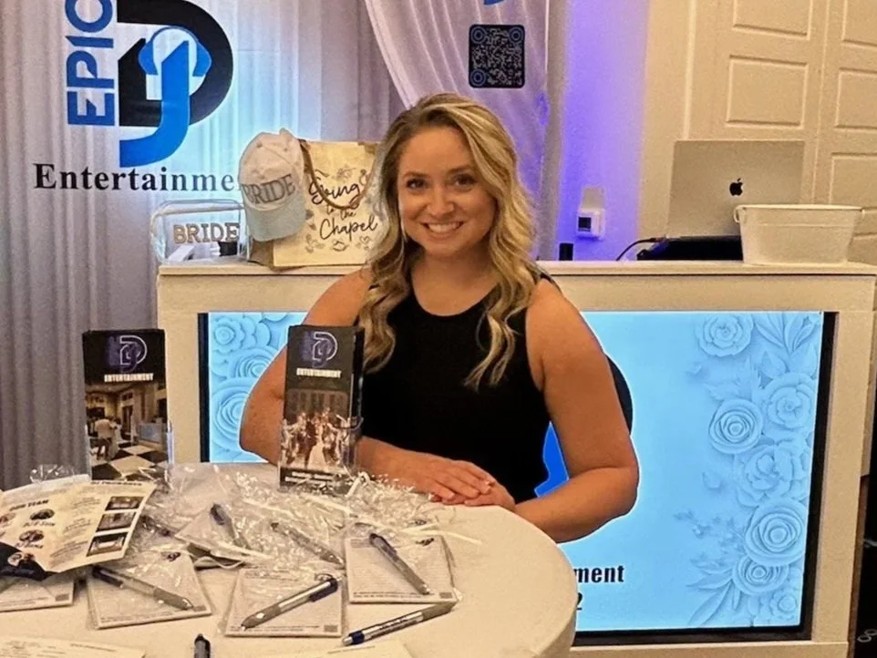 A smiling woman with long blonde hair sitting at a table with pens and papers, in front of a decorated booth with the logos "EPIC" and "Entertainment". There are promotional materials on the table and a backdrop with signs and a QR code.