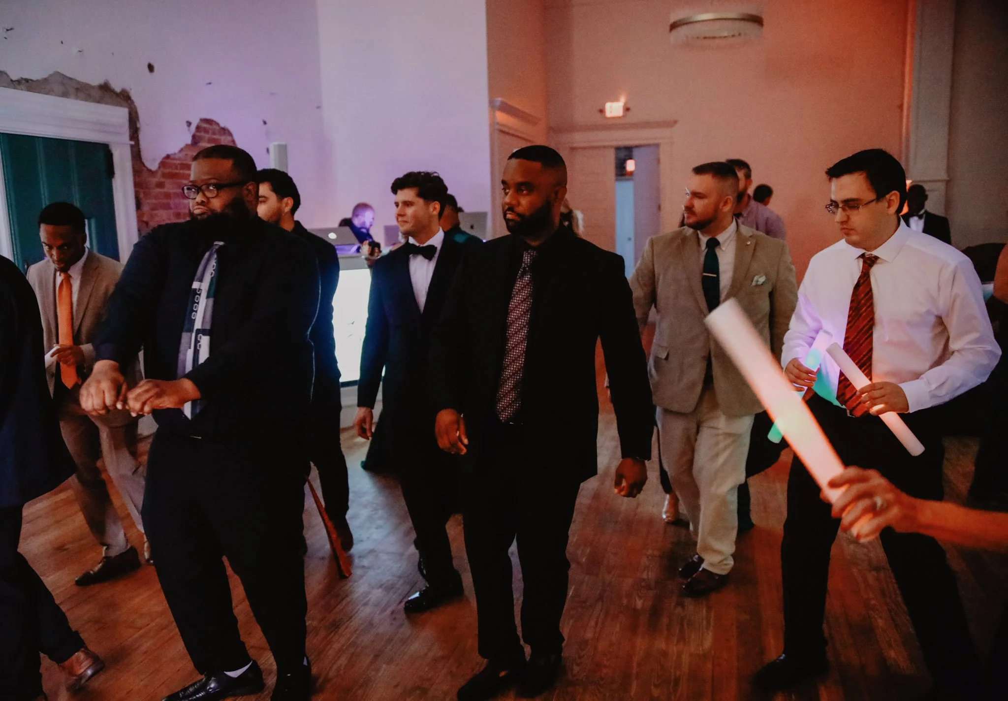Group of men in suits and formal attire walking in a dimly lit indoor setting, possibly at a formal event or party.