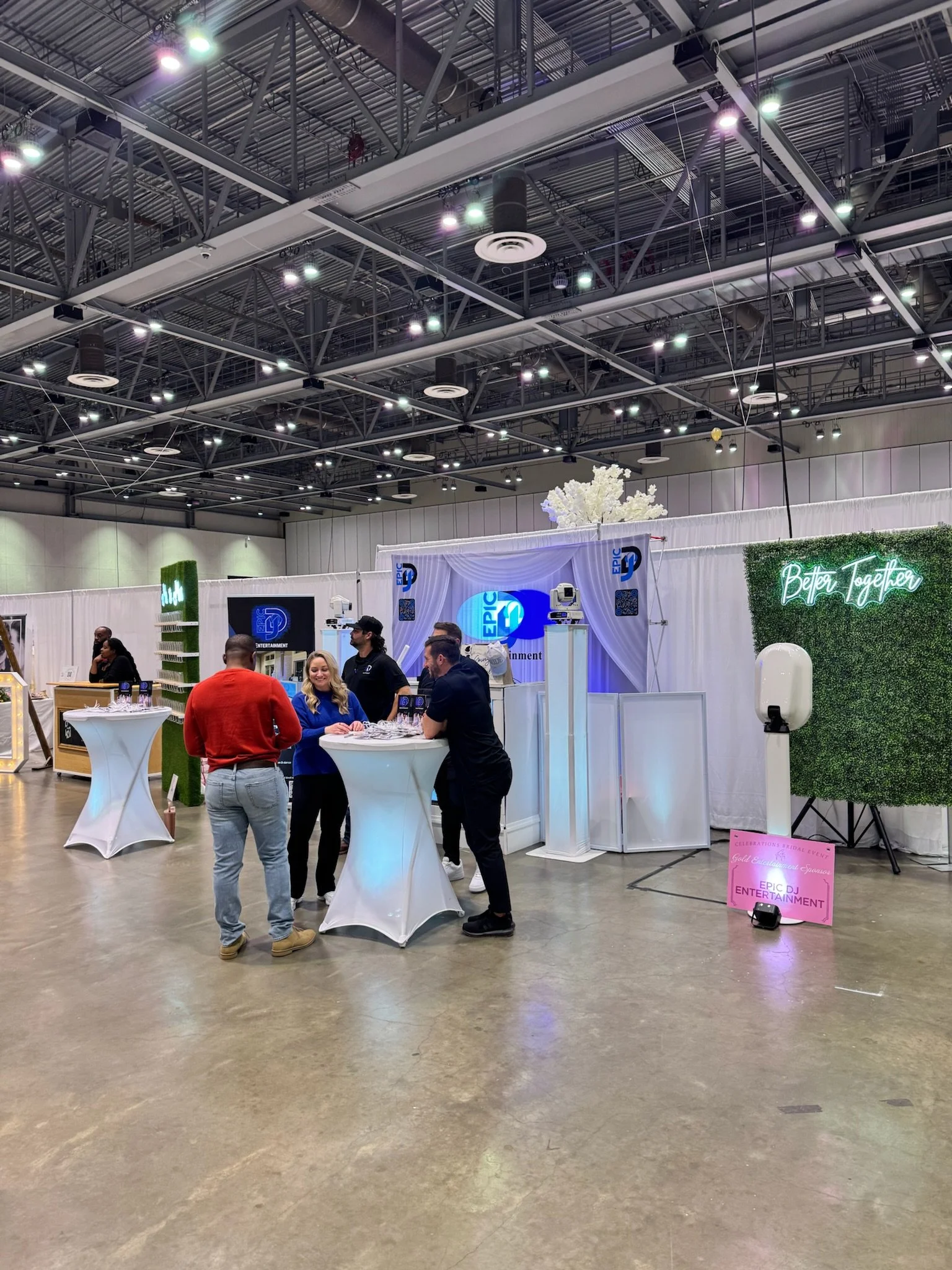 People gathered around a high table at an indoor event or convention, with a booth or display behind them featuring signage that reads 'Epic Entertainment' and 'Better Together,' decorated with greenery and neon lights.