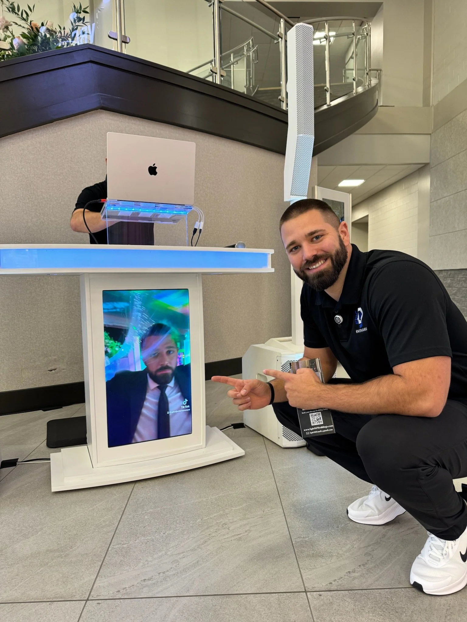 A man with a beard squatting beside a digital display shows a person in a suit taking a selfie. The man is smiling and pointing at the screen, which is showing a live selfie of himself.