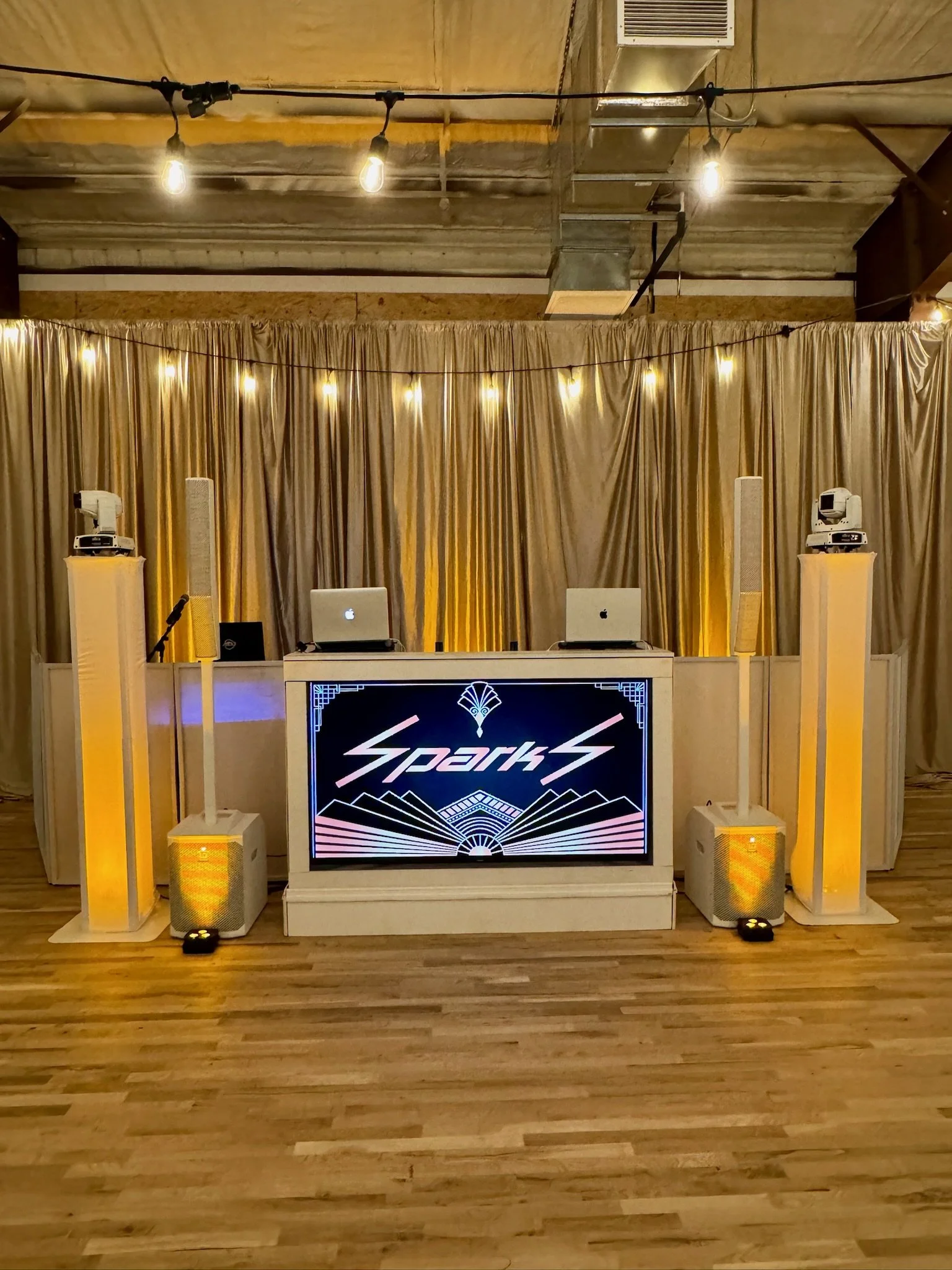 Event stage setup with a DJ booth featuring a display screen with the word 'SparkS' and an art deco design, surrounded by tall speakers, laptops, microphones, and decorative lighting with curtains and an industrial ceiling.