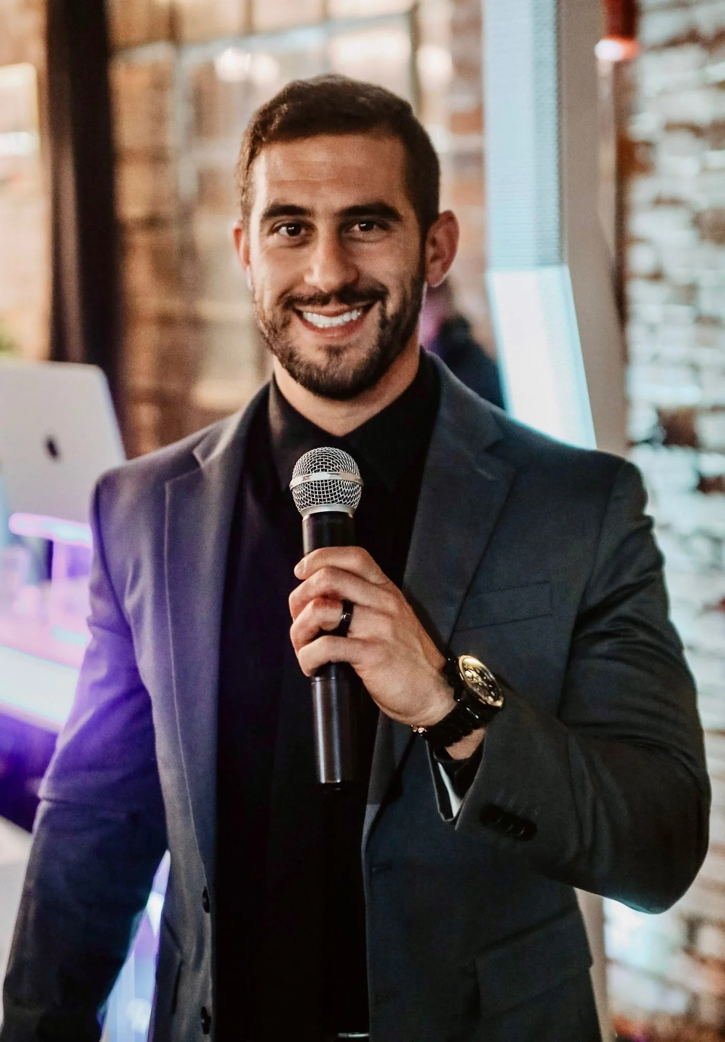 A man in a dark suit holding a microphone, smiling at an indoor event.