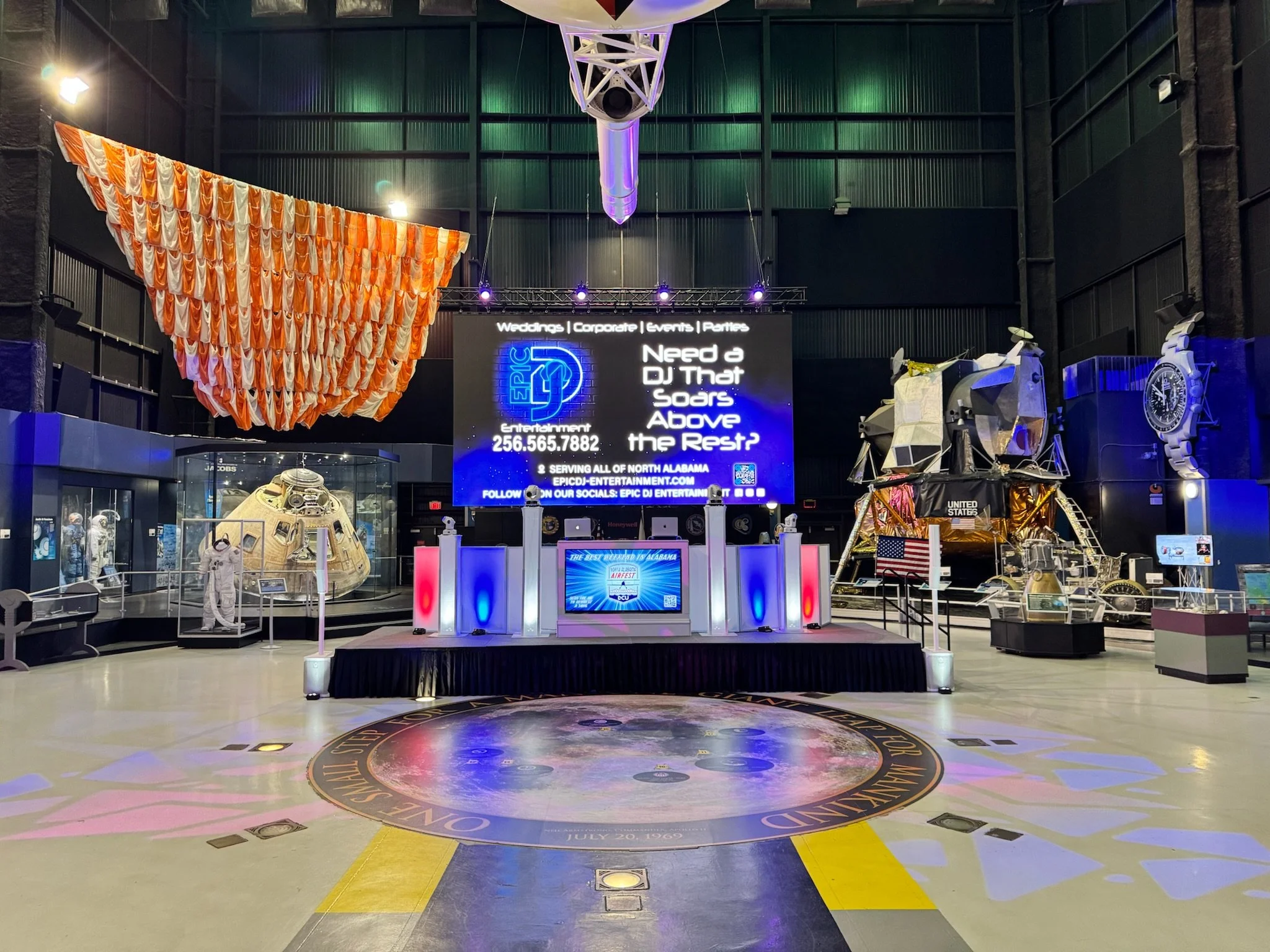 Museum exhibit featuring space exploration, including a lunar module, astronaut suit, and a mockup of a spacecraft, with a DJ stage and digital screen in a large, industrial-style space.