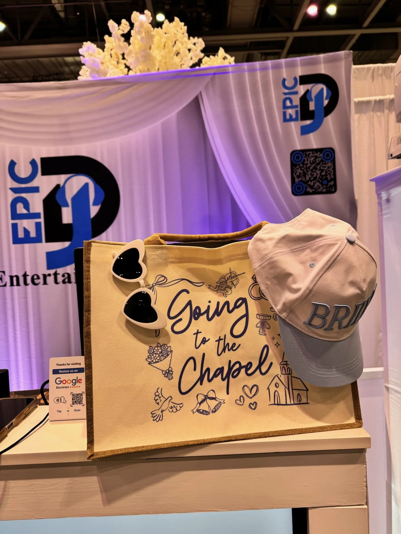 A tote bag with colorful designs and the words "Going to the Chapel" on it, hanging on a table. A pair of white sunglasses with dark lenses and a beige cap with "BR" on it are also on the table.