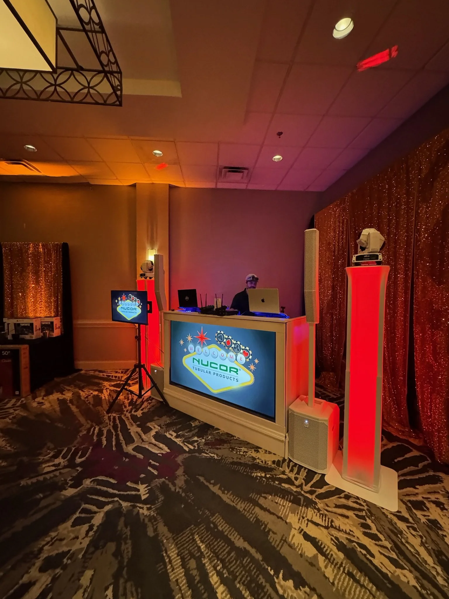 DJ booth set up with two laptop computers and audio equipment at an event with a colorful sign reading 'Welcome Nucar Tubular Products', gold sequin curtain backdrop, and patterned carpet.