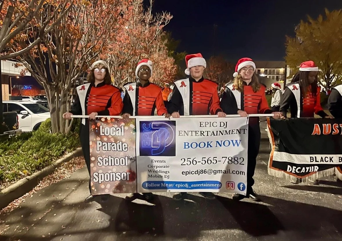 A group of children wearing band uniforms, Santa hats, and glasses, marching in a parade at night, holding a banner promoting EPIC DJ ENTERTAINMENT with contact information and social media icons.