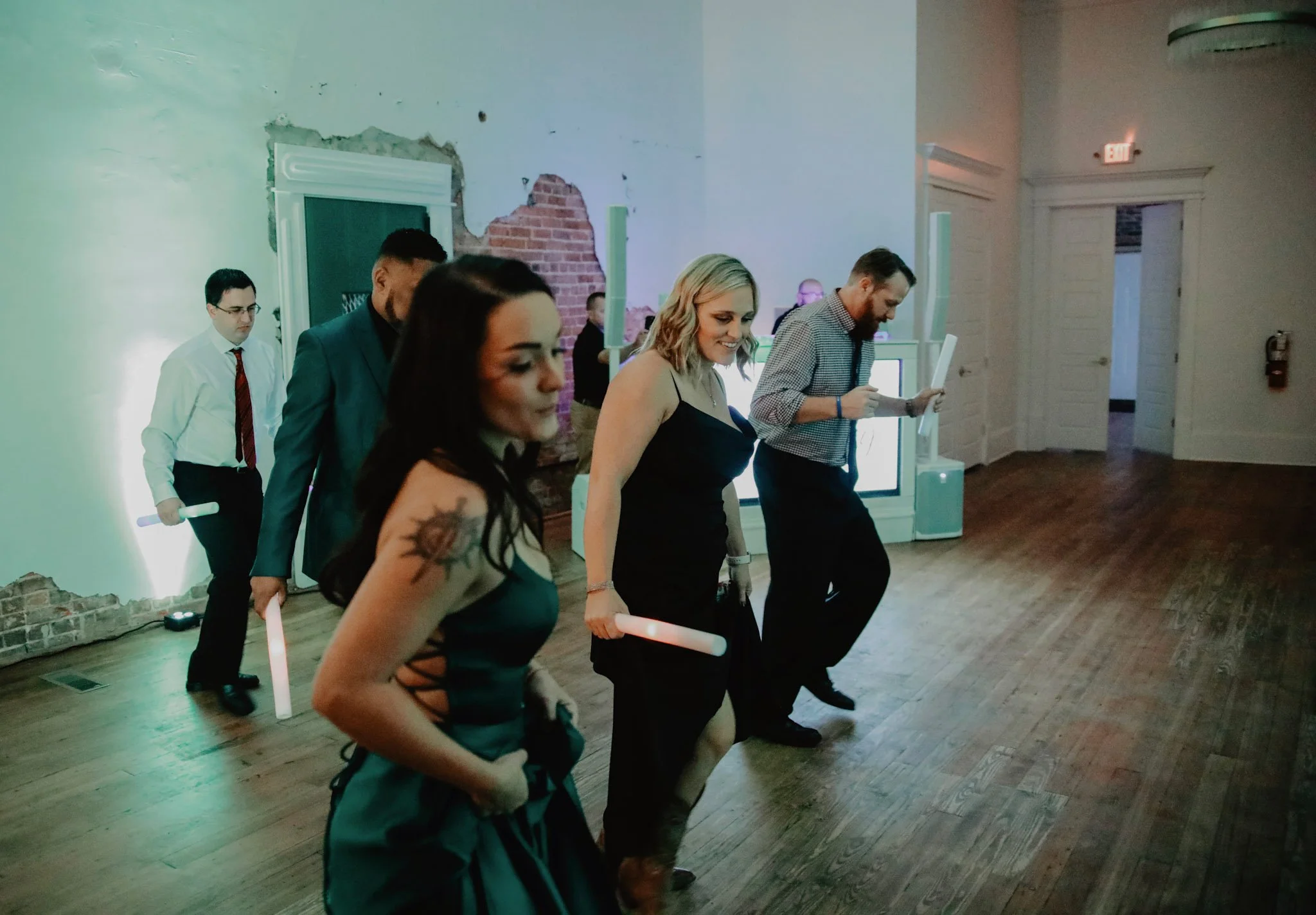 Group of people dancing with glow sticks in an indoor event space with exposed brick and peeling paint walls.
