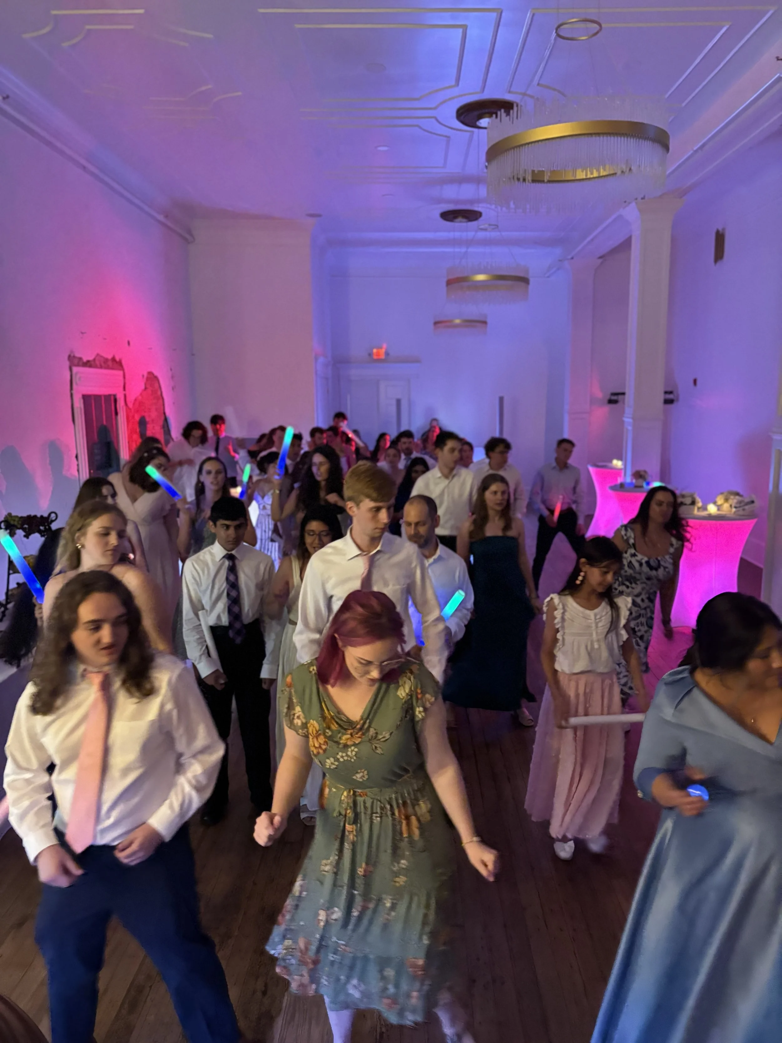 People dancing at a party or celebration in a large, decorated room with purple lighting and ornate ceiling fixtures.