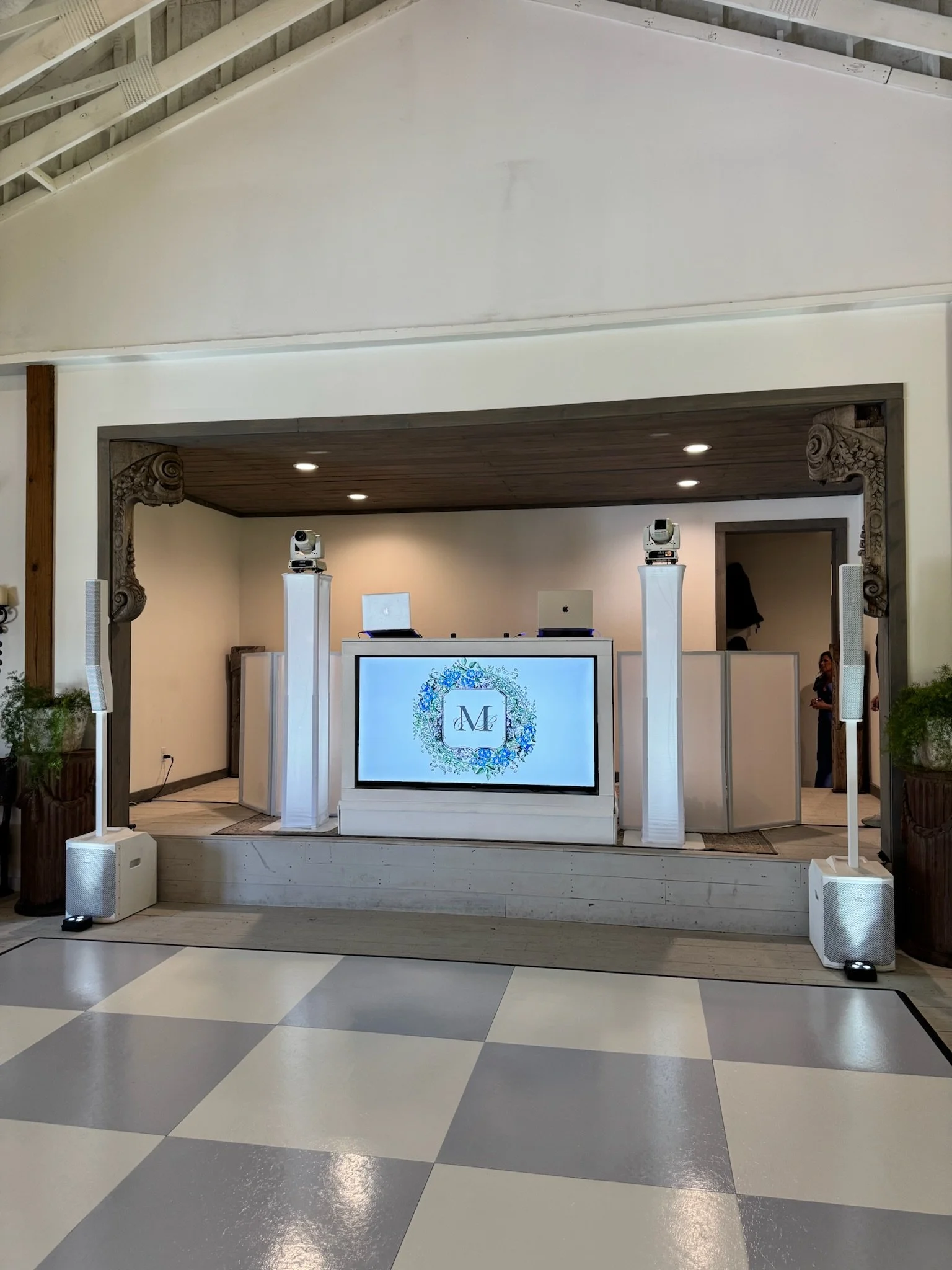 Event DJ setup with a large screen displaying a floral logo and the initials "M" and "C," speakers, laptops on DJ booths, and cameras on top of tall white stands in an indoor venue with checkered flooring.