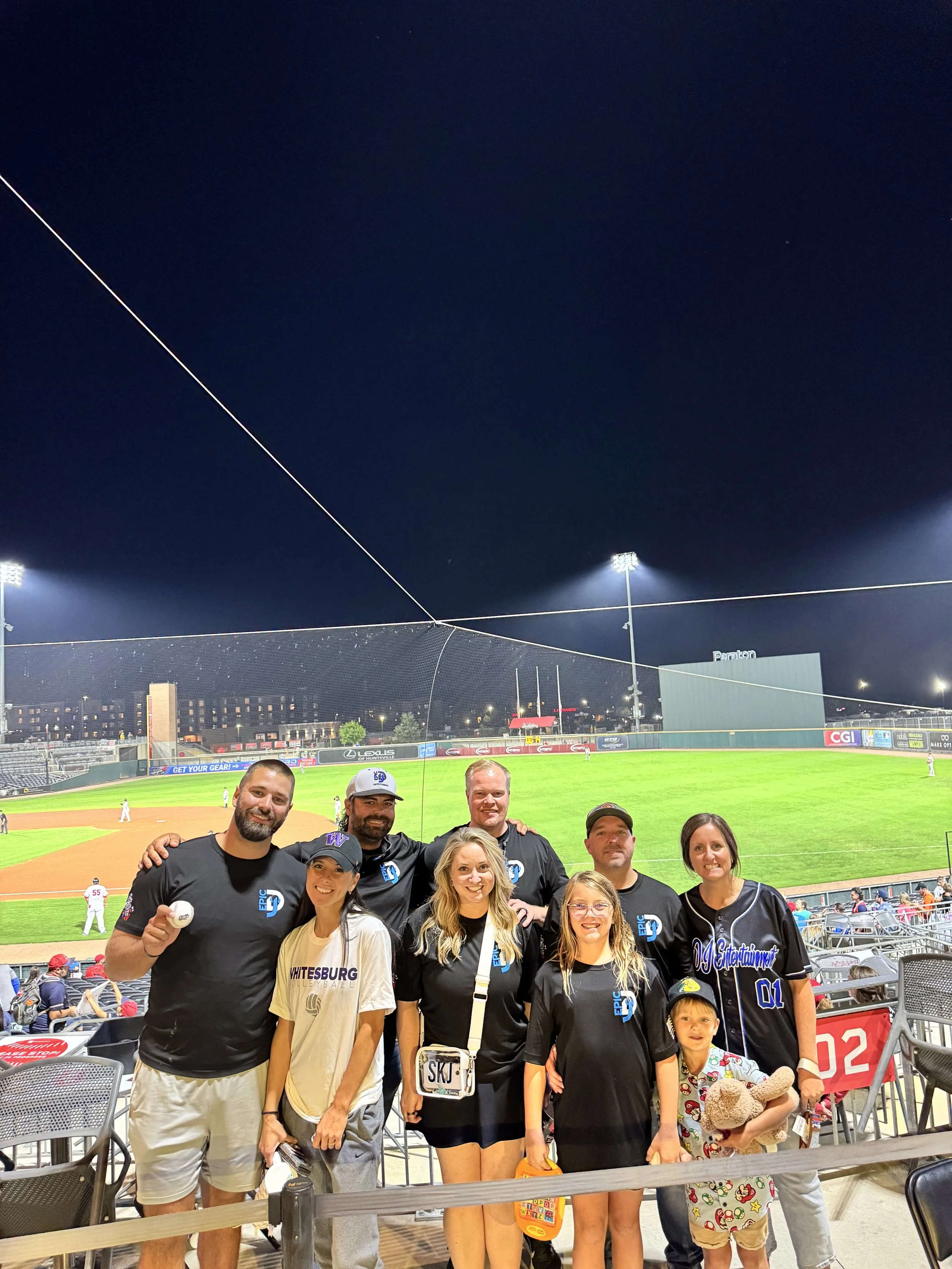 Group of people at a baseball game posing for a photo in the stands at night with the baseball field and players in the background.