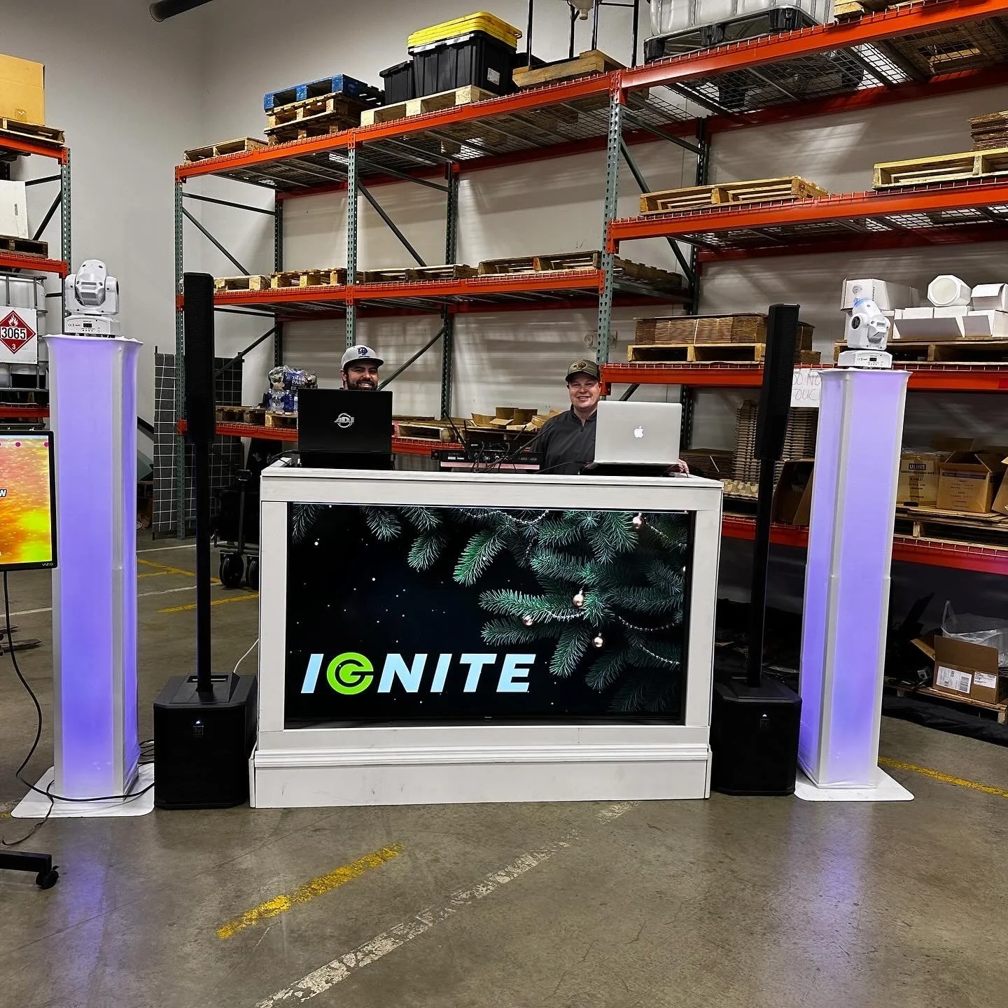 A DJ booth set up in a warehouse with a large screen displaying the word 'IGNITE' and a pine branch graphic. Two DJs are behind the booth, one with a black laptop and the other with a silver MacBook. There are speakers and lighting equipment on tall 