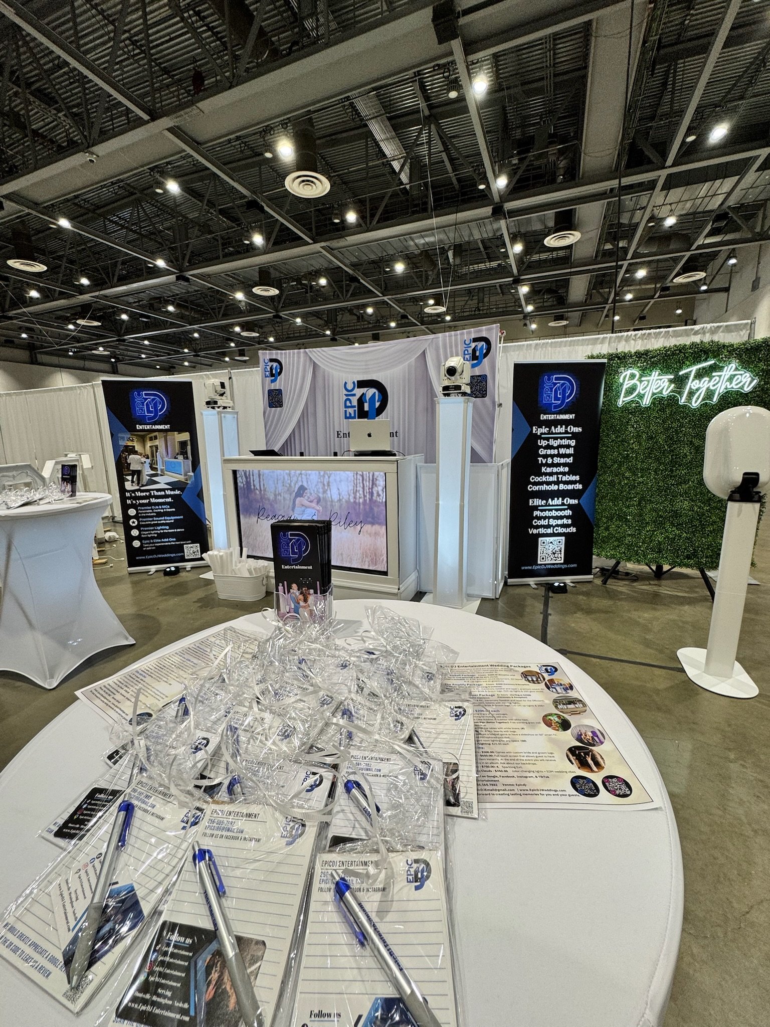 Exhibition booth with banners, a table with promotional materials, and a green wall with neon text that reads 'Better Together' inside a large event hall.