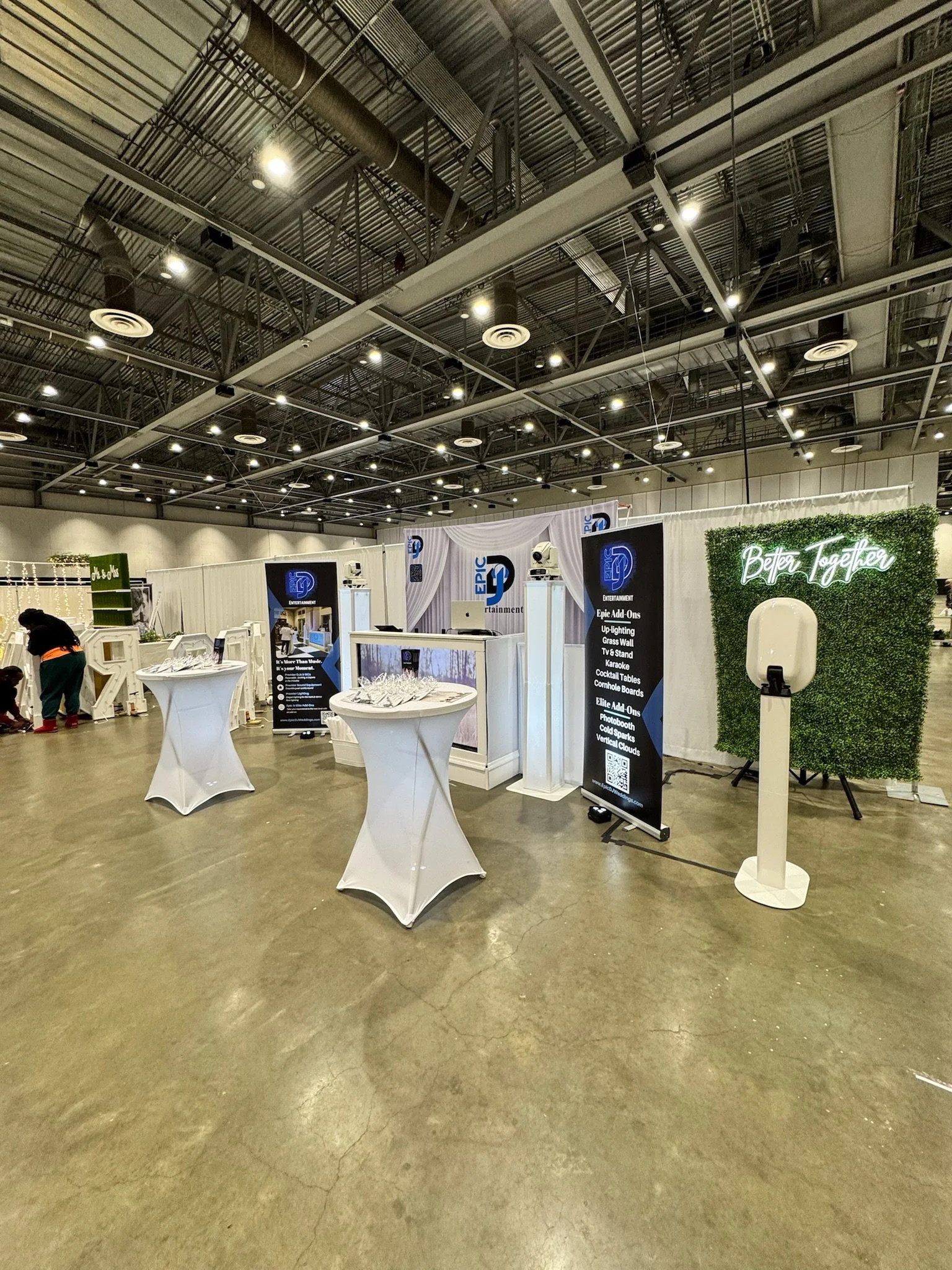 Exhibition booth with banners and tables in a large indoor venue, featuring a neon sign that reads 'Better Together' and a green hedge wall.