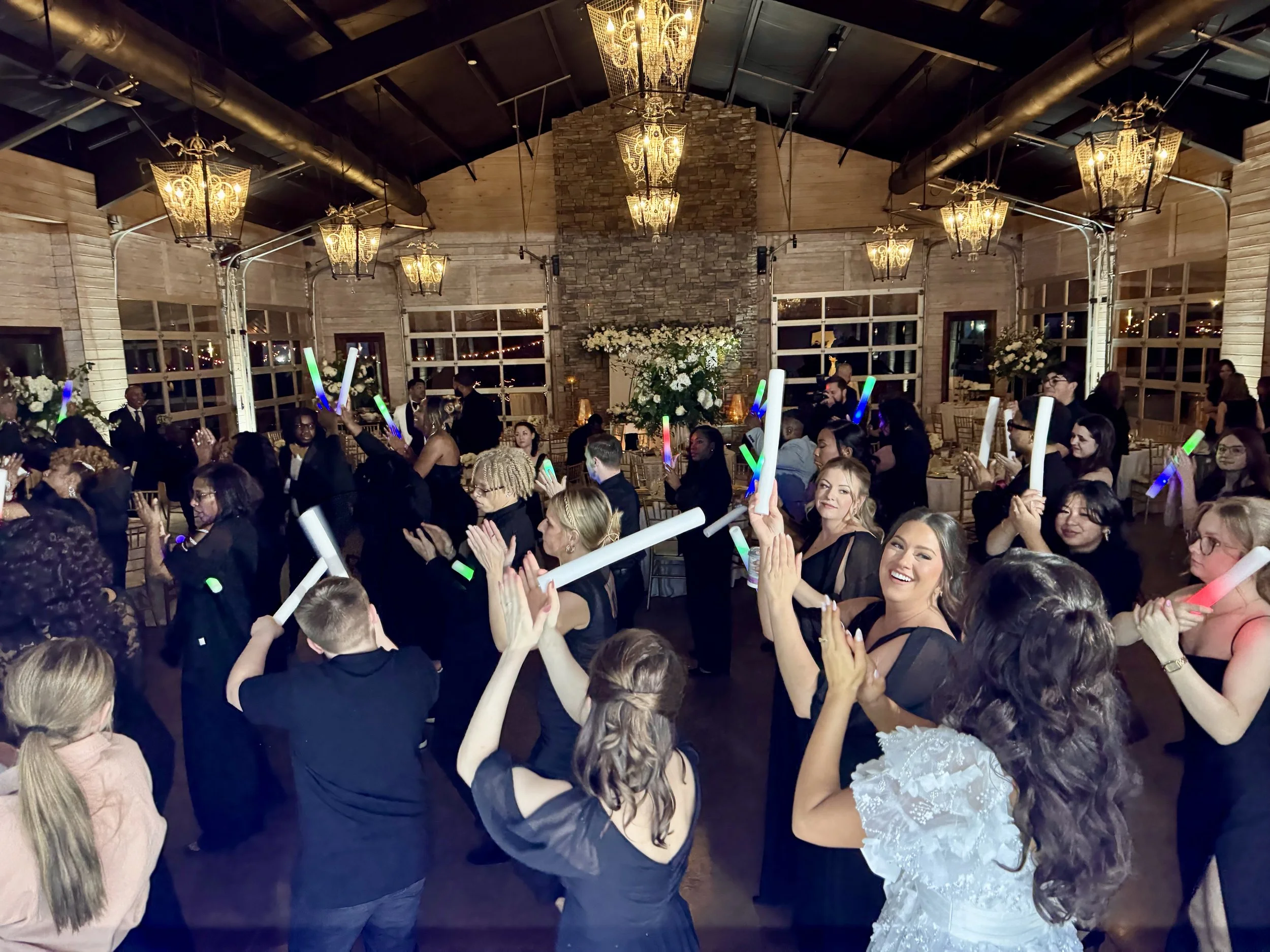 People dancing and celebrating at a party with glowing foam sticks in a decorated hall with chandeliers and floral arrangements.