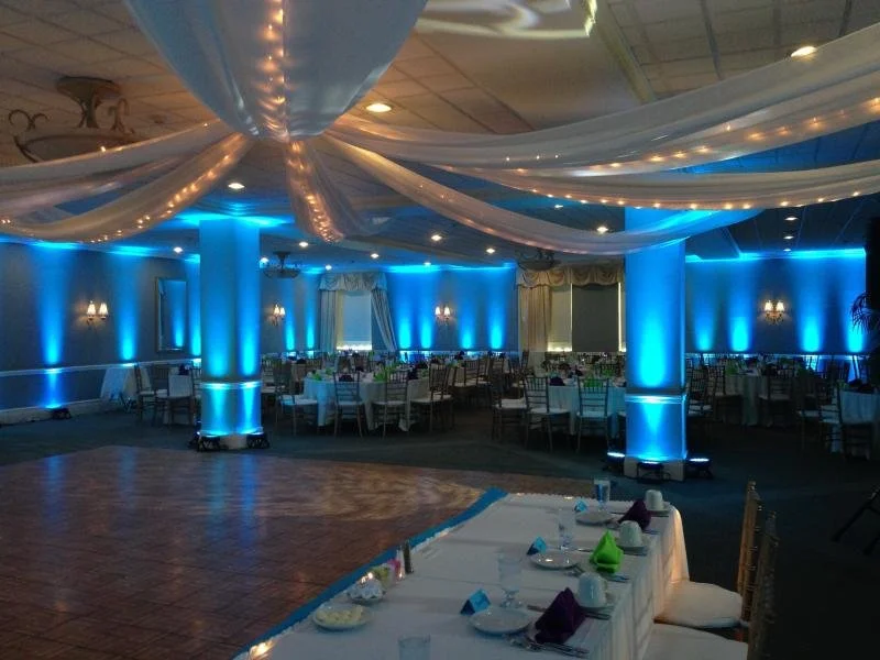 An event hall decorated with white drapes and blue lighting, featuring tables set for a celebration.