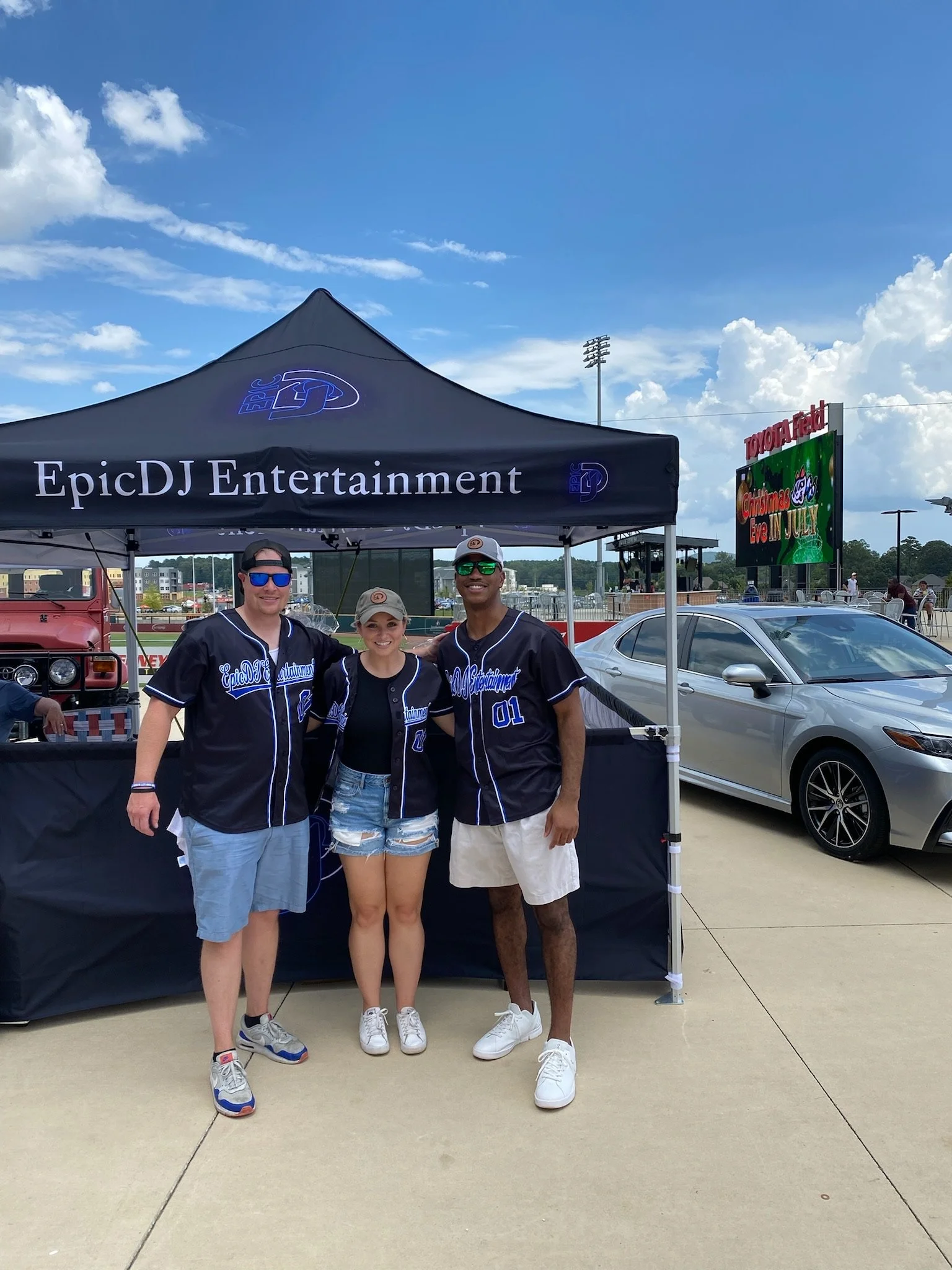 Three people standing together in front of a tent that says Epic DJ Entertainment at a sports event, with a parking lot and a digital sign and a silver car in the background.