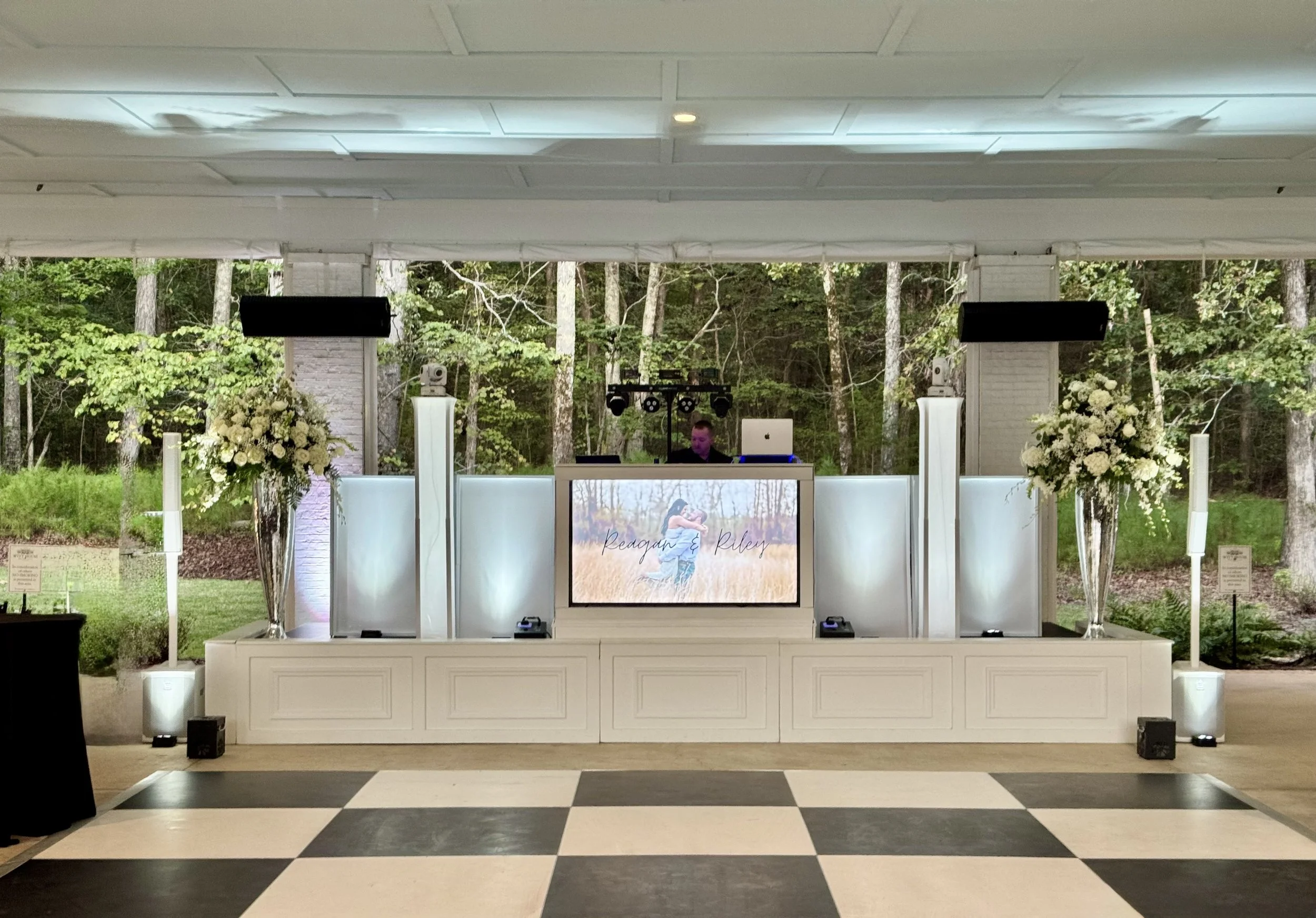 Wedding reception setup with a DJ booth, large flower arrangements, and a black and white checkered dance floor, outdoors in a wooded area.